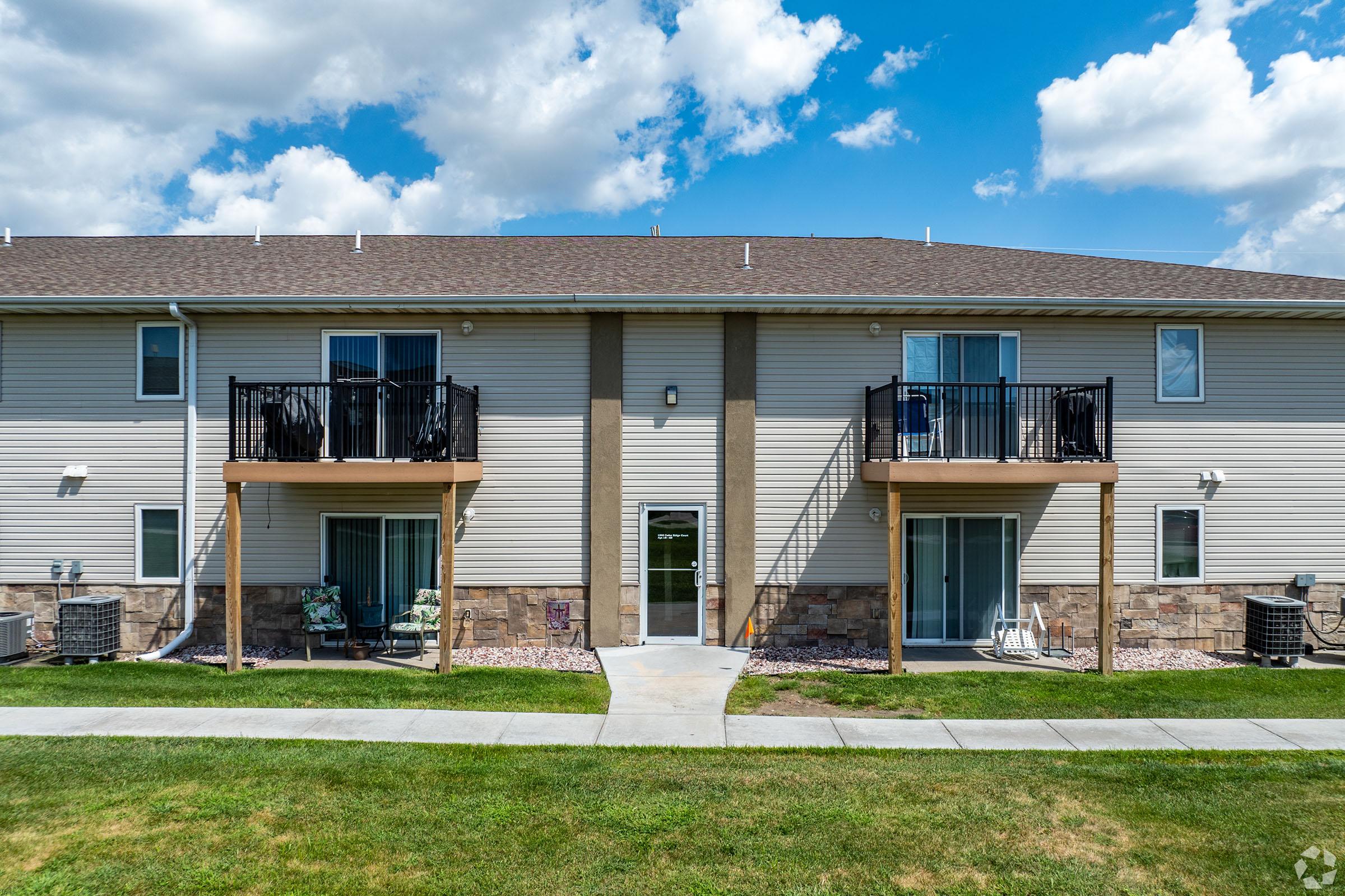Two-story apartment building with beige siding, featuring two balconies on the ground floor and large windows. A concrete pathway leads to the entrance, bordered by a well-maintained grassy area. The sky is partly cloudy, adding a vibrant feel to the scene.