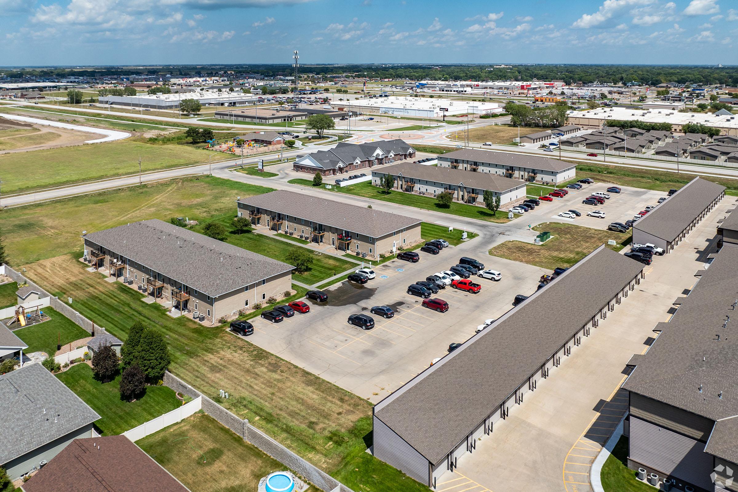 Aerial view of a residential area featuring multiple apartment buildings with parked cars. Open green spaces and a nearby road are visible, along with commercial buildings in the background. The scene is set under a clear blue sky with scattered clouds.