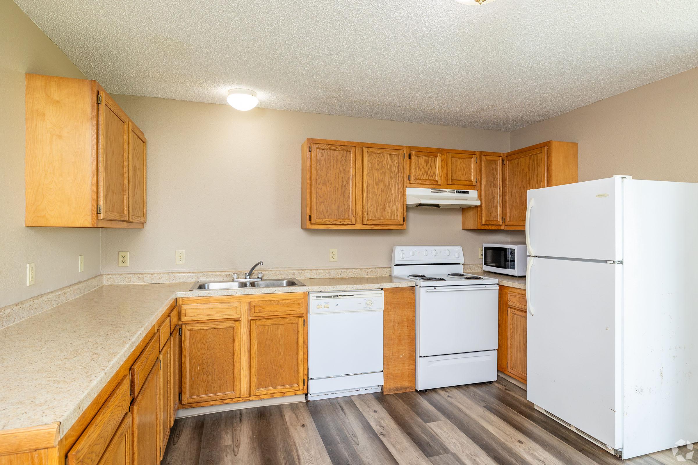 A modern kitchen featuring light wooden cabinets, a double sink, a white stove, and a microwave. It includes a dishwasher and a large white refrigerator. The countertop is a light color, and the flooring is wood laminate. Soft lighting illuminates the space, giving it a warm and inviting atmosphere.