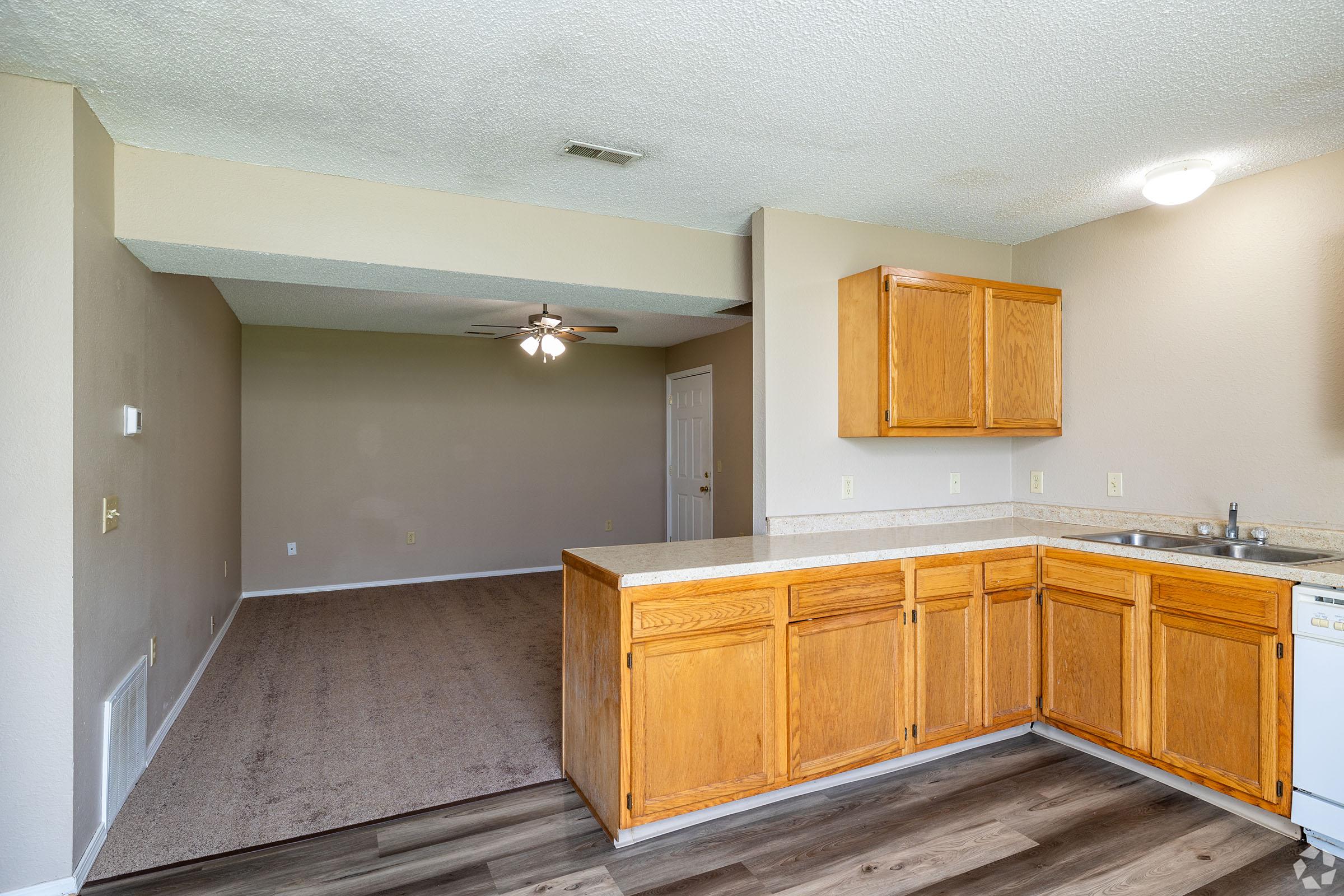 A spacious kitchen with wooden cabinets, a sink, and dishwasher. The kitchen area opens to a living space featuring neutral-colored walls and carpet. A ceiling fan is visible in the living area, which is well-lit with natural daylight.