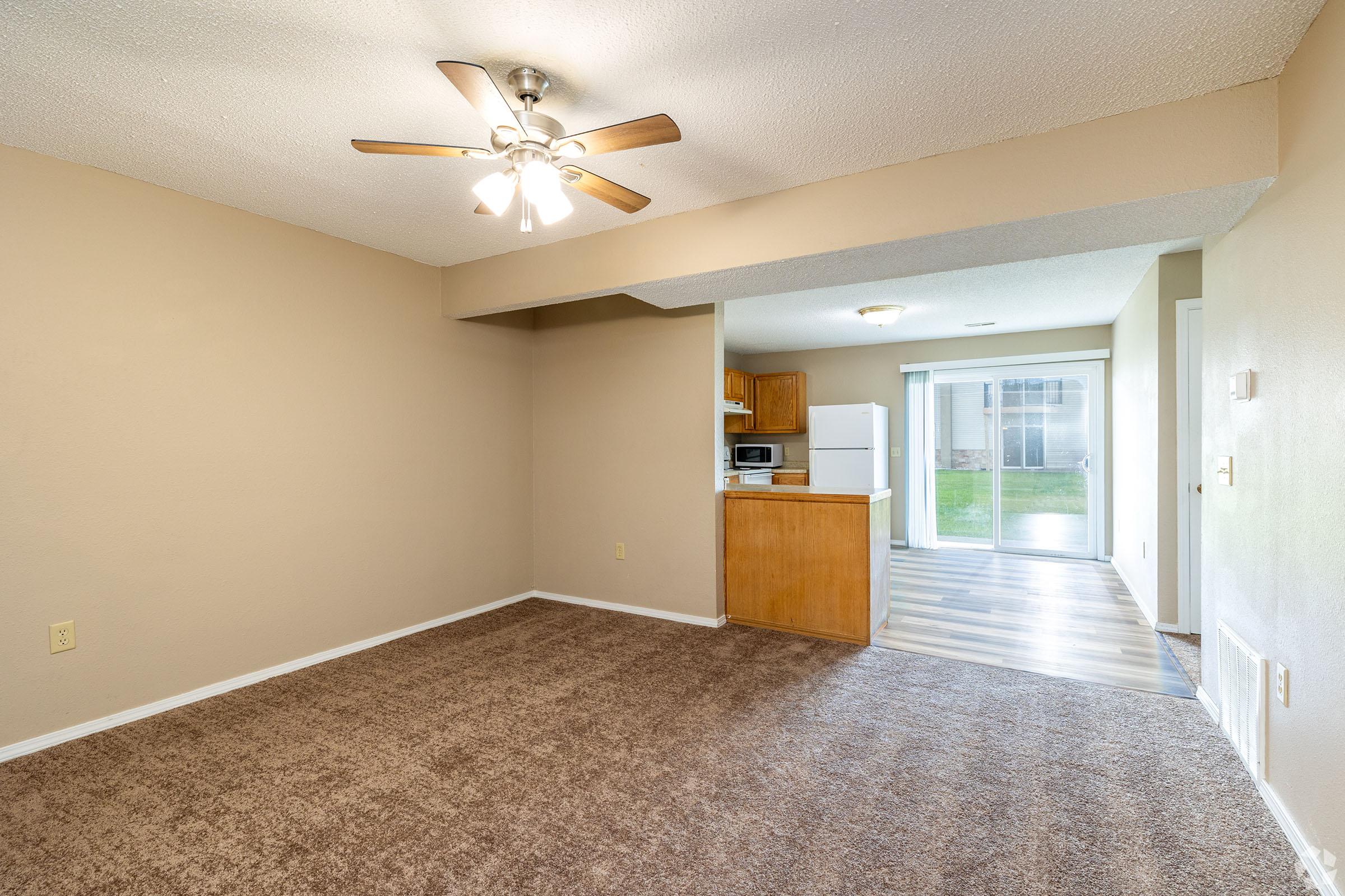 A spacious living area with light brown carpet, featuring a ceiling fan and an open layout. To the left, a kitchen is visible with wooden cabinets and appliances. A large window in the background allows natural light and shows a grassy outdoor area.
