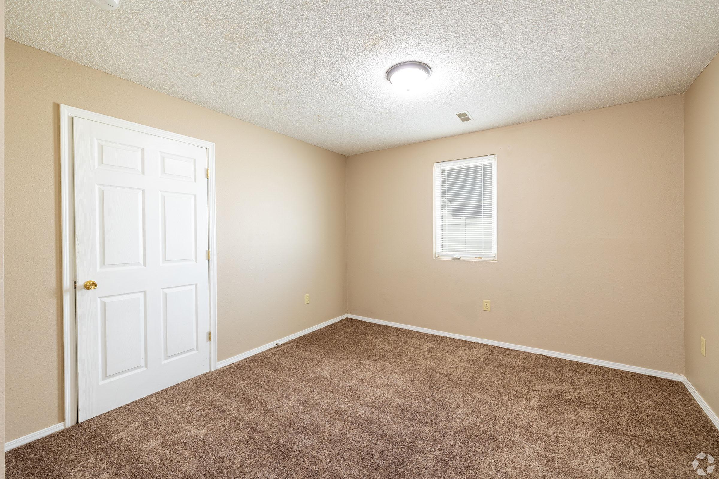 A small, empty room with light brown walls and carpet. There is a white door on the left and a window with blinds on the right. The ceiling has a simple light fixture. The overall appearance is clean and well-maintained, suitable for various purposes like an office or bedroom.