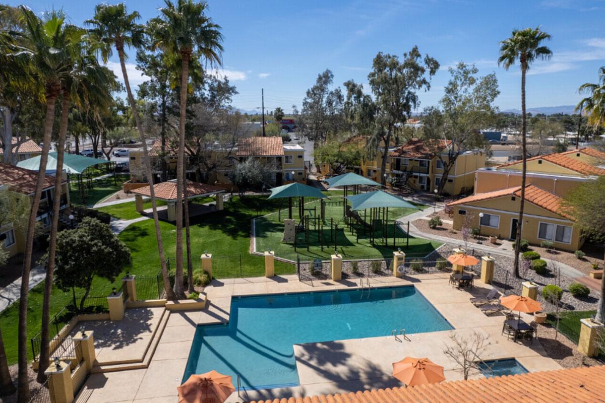 Aerial view of a resort-style pool area with lounge chairs and umbrellas, surrounded by landscaped gardens and palm trees. Nearby, there is a grassy play area with playground equipment. The setting includes buildings with tiled roofs and a clear blue sky in the background.