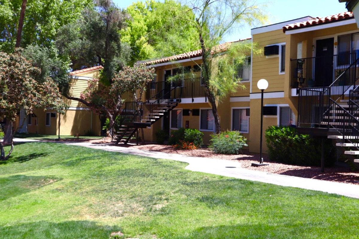 A view of a landscaped courtyard featuring two-story apartment buildings with yellow exteriors. The buildings have balconies and outdoor staircases. Green grass and shrubs surround the pathways, with trees providing shade. The scene conveys a tidy, residential atmosphere.