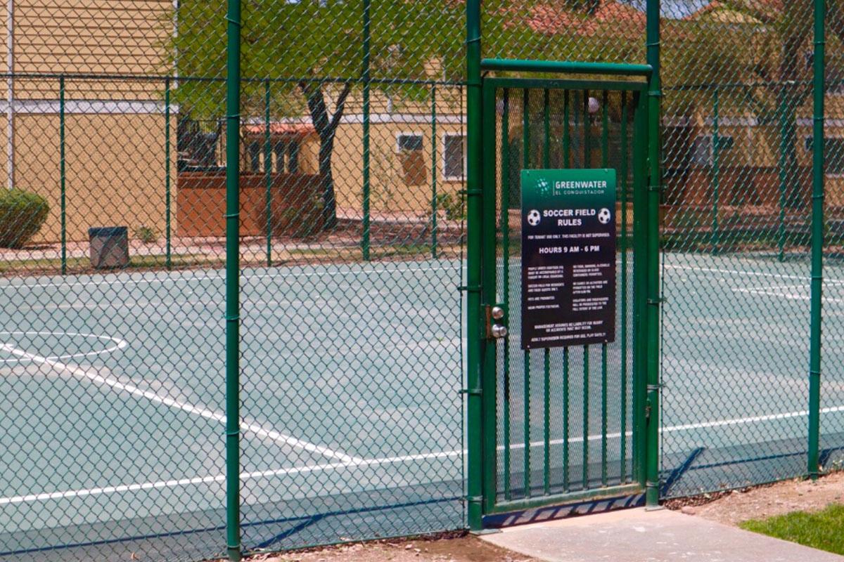 A green fenced soccer field with a locked gate, featuring a sign detailing the rules and hours of use. The field is empty with trees and buildings visible in the background, indicating a recreational area.