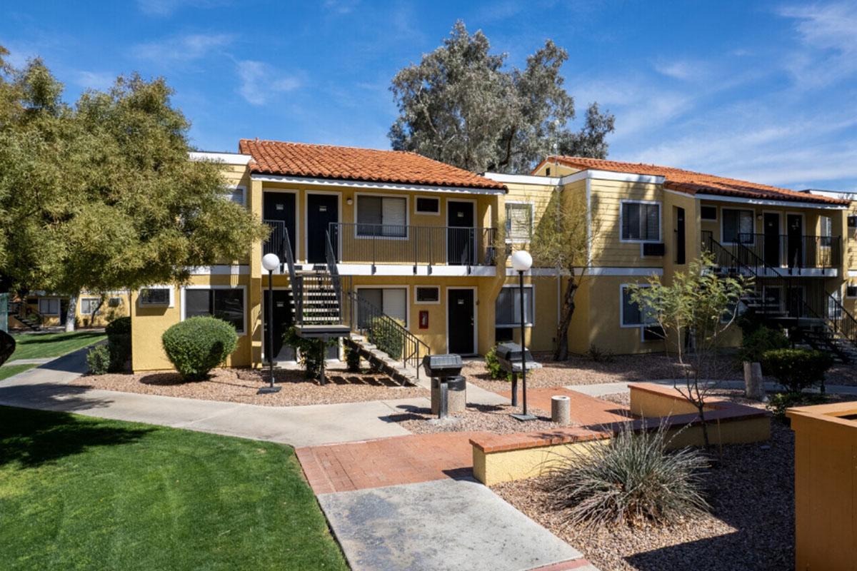 A view of an apartment complex featuring two-story buildings with yellow exteriors and terracotta roof tiles. The area includes landscaped gardens, pathways, and outdoor grills, with palm trees and shrubs enhancing the scenery under a clear blue sky.