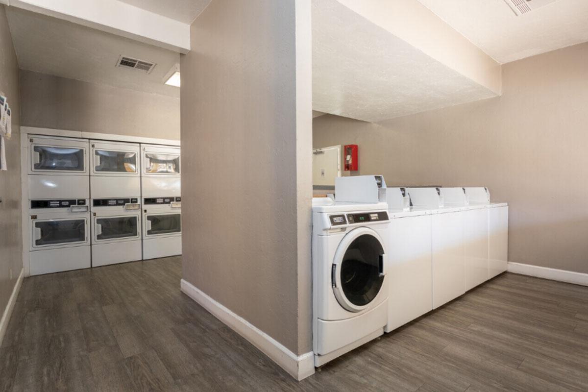 A clean and well-lit laundry room featuring multiple white washing machines and dryers. The walls are painted a neutral color, and the flooring is a light wood finish. There is a fire extinguisher mounted on the wall, and the space appears organized and functional, with various laundry appliances available for use.