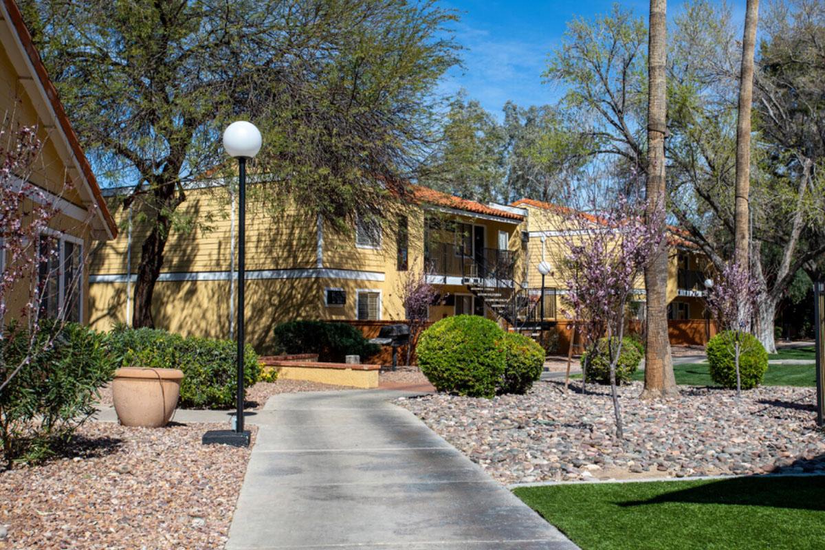 A pathway leads through a landscaped area with shrubs and ornamental trees, flanked by yellow apartment buildings. A lamp post stands beside the walkway, and the scene features greenery and pebbles, capturing a sunny, tranquil residential environment.