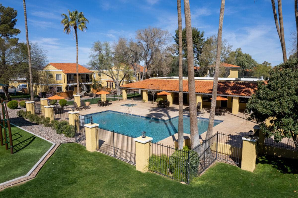A tranquil outdoor pool area surrounded by palm trees and lush landscaping. The pool is enclosed by a decorative fence and features lounge chairs and umbrellas. Nearby, there are two-story buildings with terracotta roofs, adding to the serene atmosphere. Clear blue skies create a warm and inviting setting.