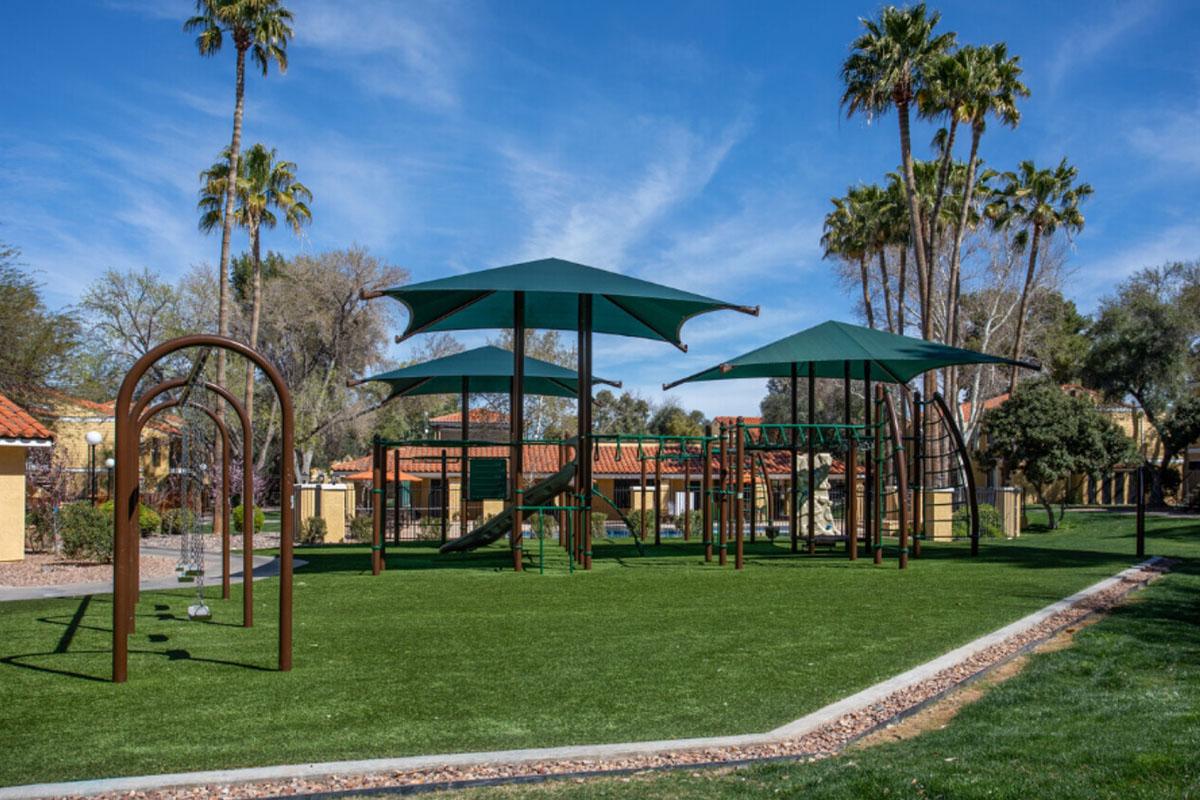A playground featuring various climbing structures and swings, shaded by large green umbrellas. Surrounding the playground is a well-maintained grassy area, with palm trees in the background under a clear blue sky.