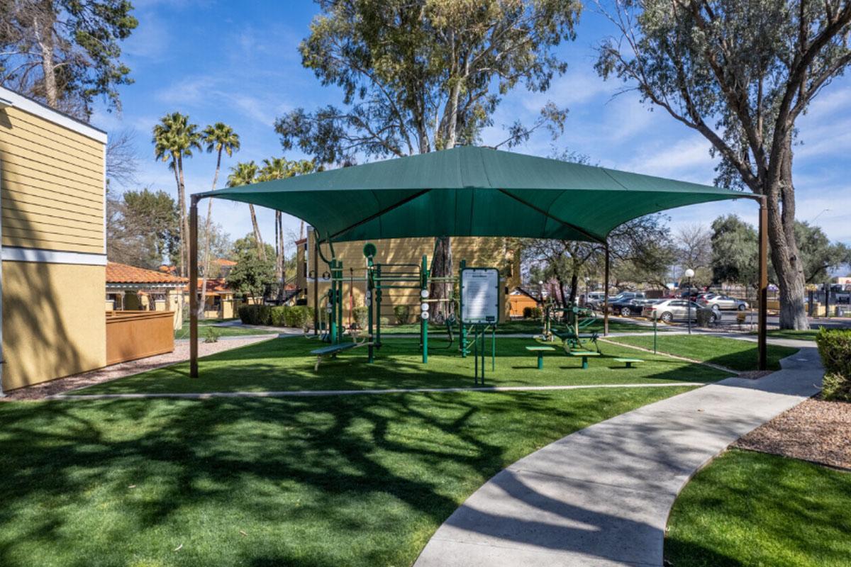 Outdoor fitness area covered by a large green canopy, featuring workout equipment on a grassy surface. Surrounding trees and a pathway leading through the space, with nearby buildings and parked cars visible in the background. Clear blue sky above adds to the inviting atmosphere.