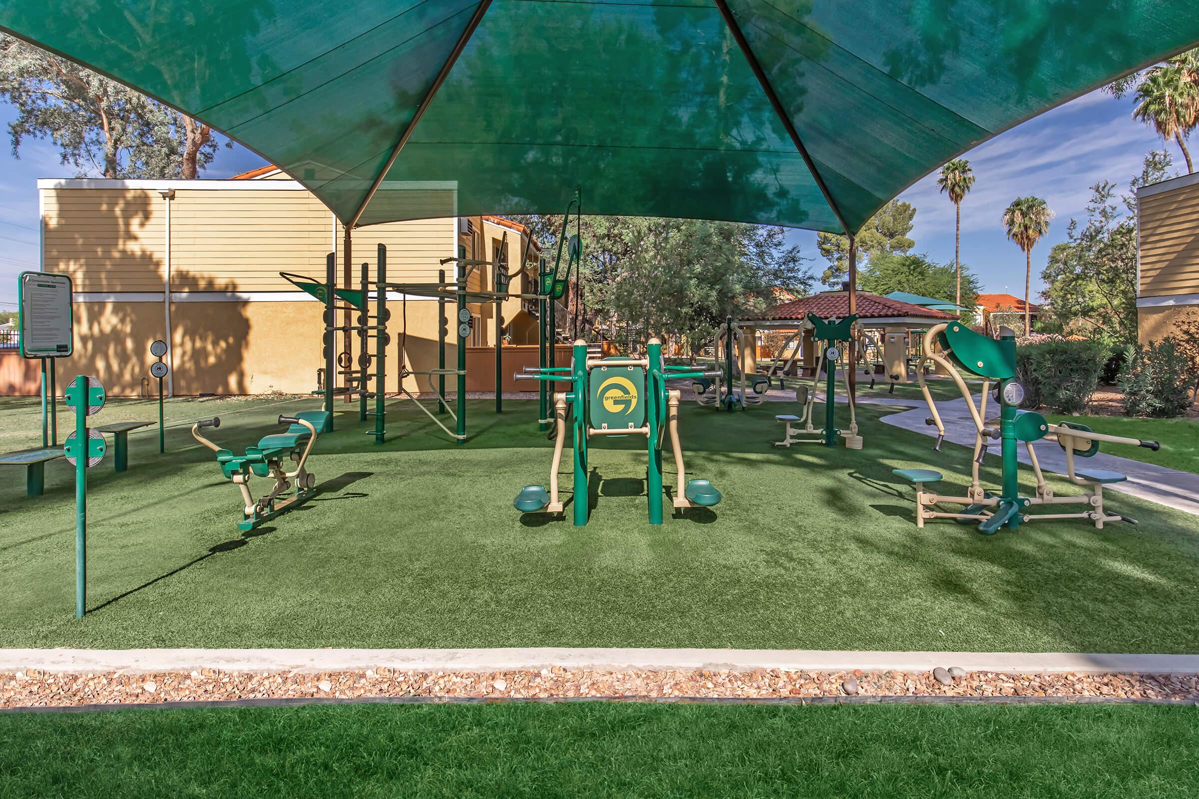 Outdoor fitness area with green exercise equipment covered by a large canopy. The space features various machines for strength training and cardio, surrounded by grass and palm trees. The area is well-maintained and designed for outdoor workouts.