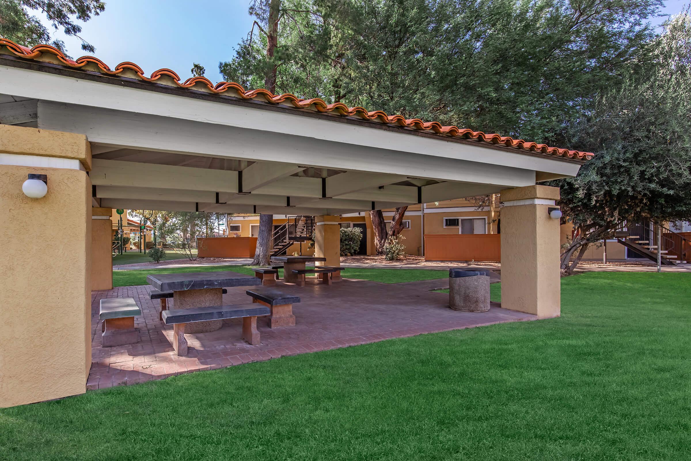 A shaded outdoor picnic area featuring a concrete table and benches under a covered structure. Lush green grass surrounds the space, with trees providing additional shade. Residential buildings can be seen in the background, creating a serene and inviting atmosphere for gatherings.