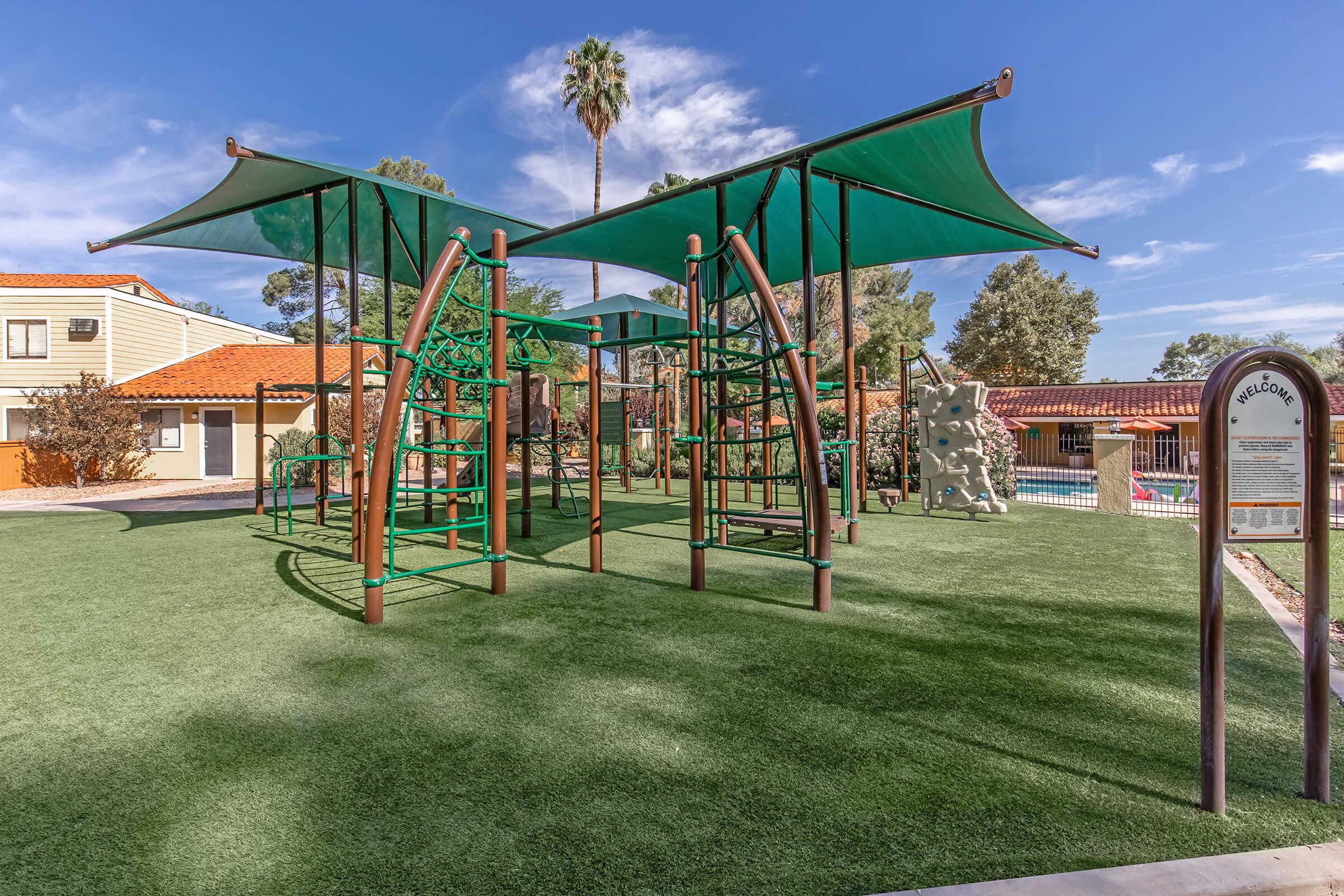 Playground featuring green climbing structures with large canopies, set on artificial grass. In the background, there are palm trees and buildings with a pool area visible. A sign marked "Welcome" can be seen nearby, indicating the entrance to the playground area.