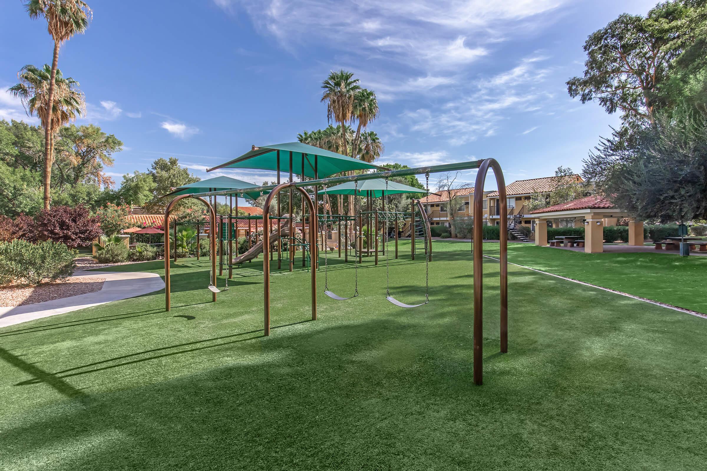 A playground featuring metallic swings with green canopies, surrounded by lush artificial grass. In the background, there are trees and buildings with a sunny sky above, creating a serene outdoor environment ideal for children to play.