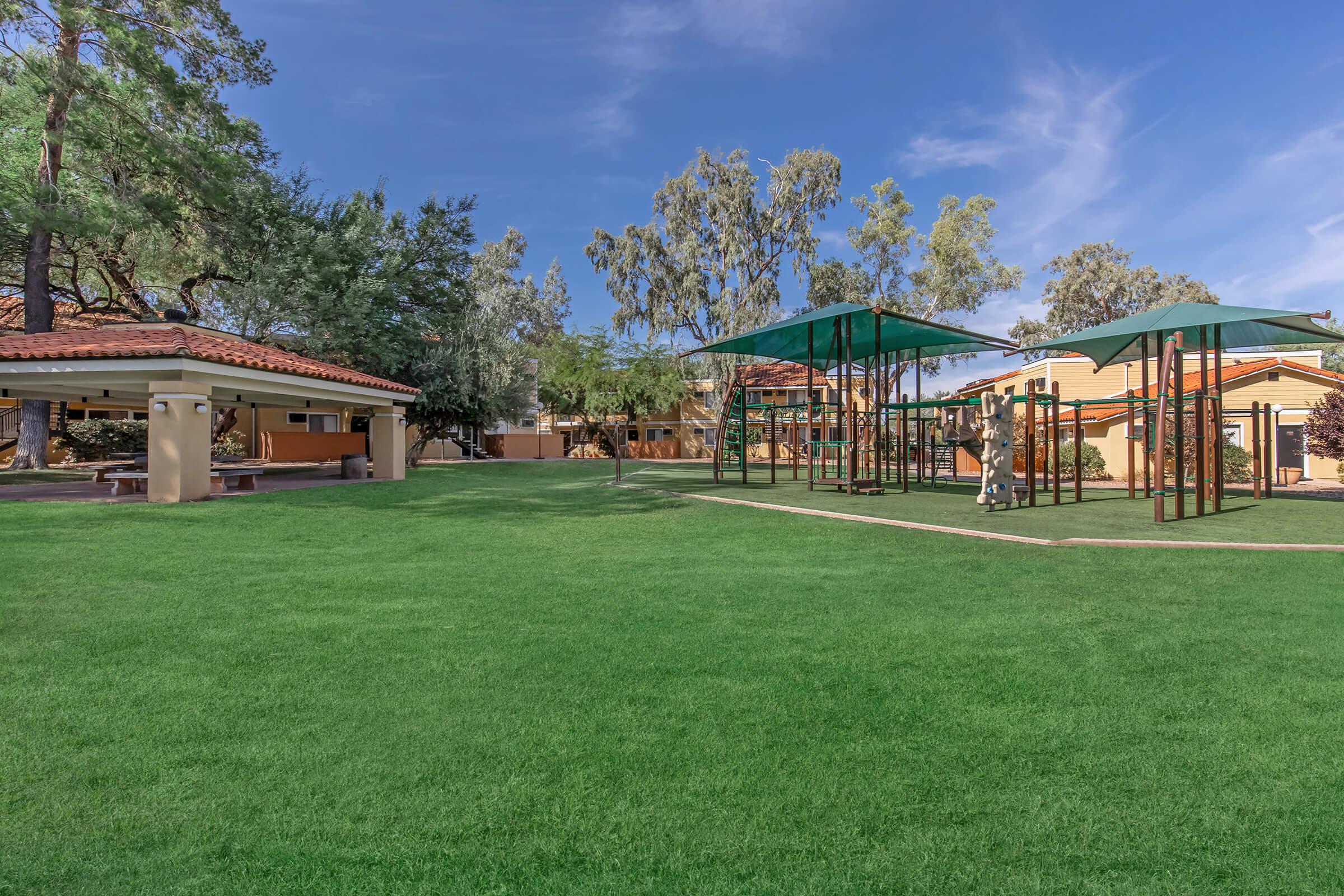 A sunny playground area featuring green grass, two large shaded structures over playground equipment, and a nearby pavilion with benches surrounded by trees and residential buildings in the background.