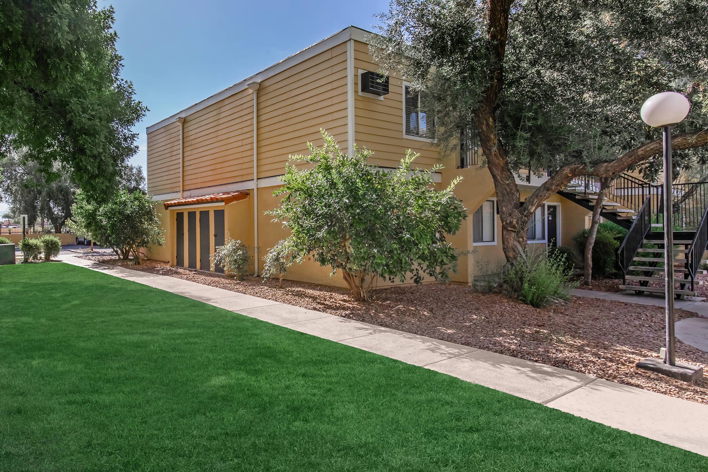 A two-story building with yellow siding surrounded by greenery, including shrubs and trees. A grassy area is in the foreground, and a path leads to the building. A staircase is visible on the right side, along with exterior lighting fixtures. The scene is bright and sunny, showcasing a well-maintained outdoor space.