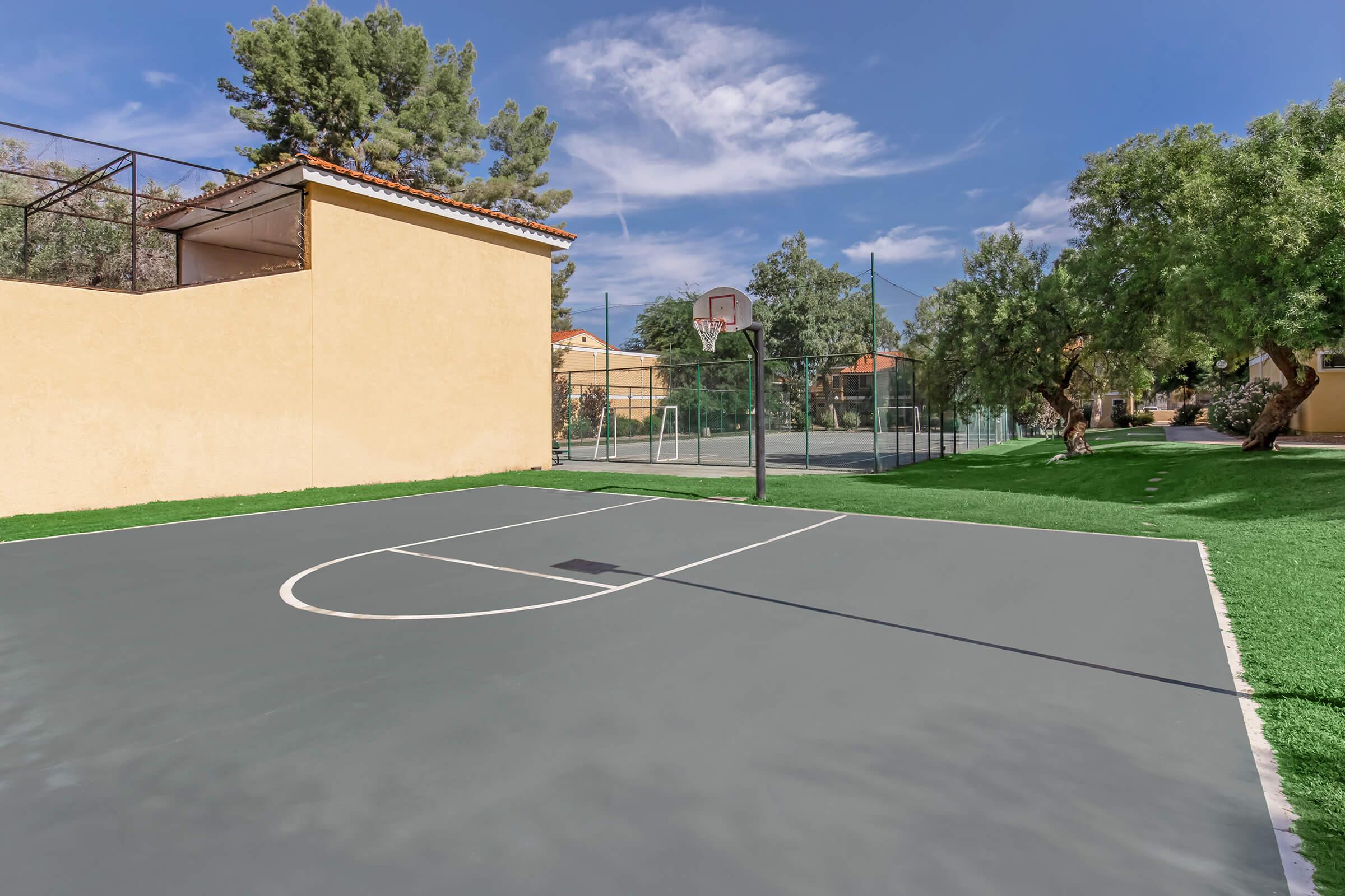A view of a basketball court featuring a gray surface with a half-court line and a hoop. In the background, there is a green tennis court visible behind a fence, surrounded by trees and a light-colored building under a clear blue sky with scattered clouds.