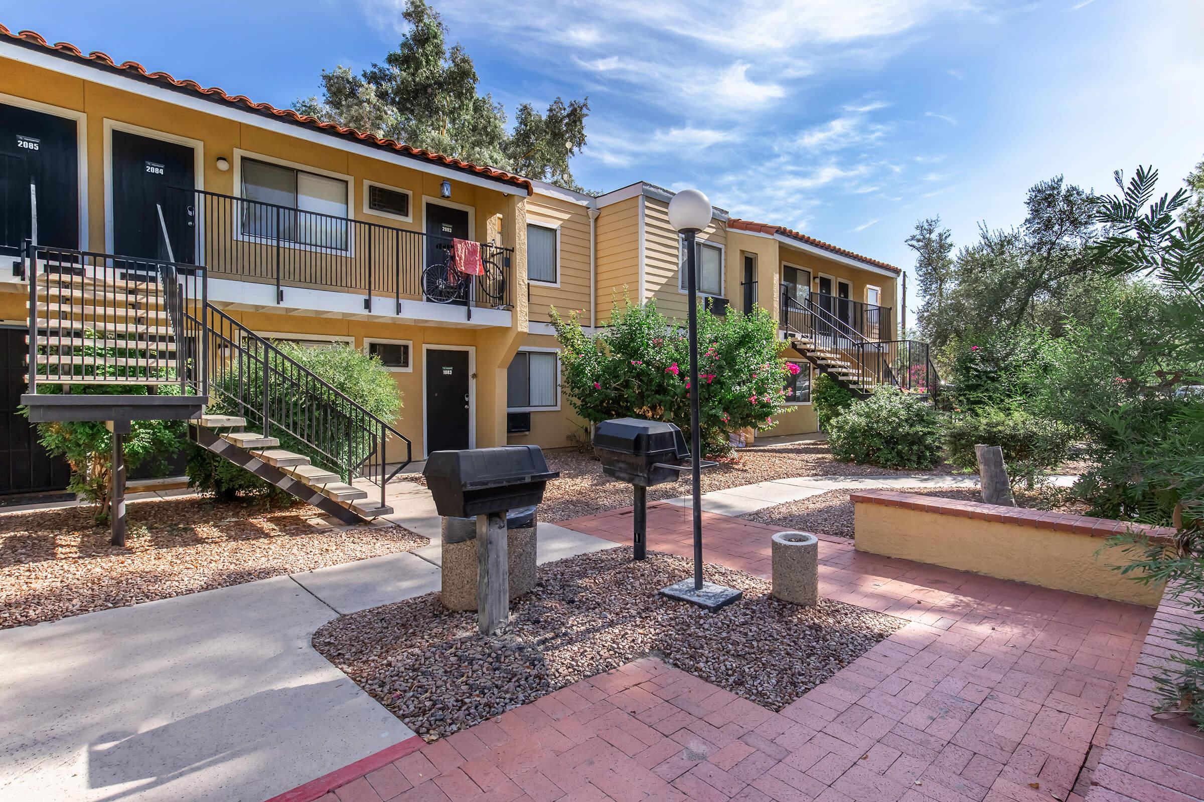 A view of an apartment complex courtyard featuring two-story buildings with staircases, several barbecue grills, and landscaped areas with plants and gravel. The sky is blue with light clouds, creating a bright atmosphere.