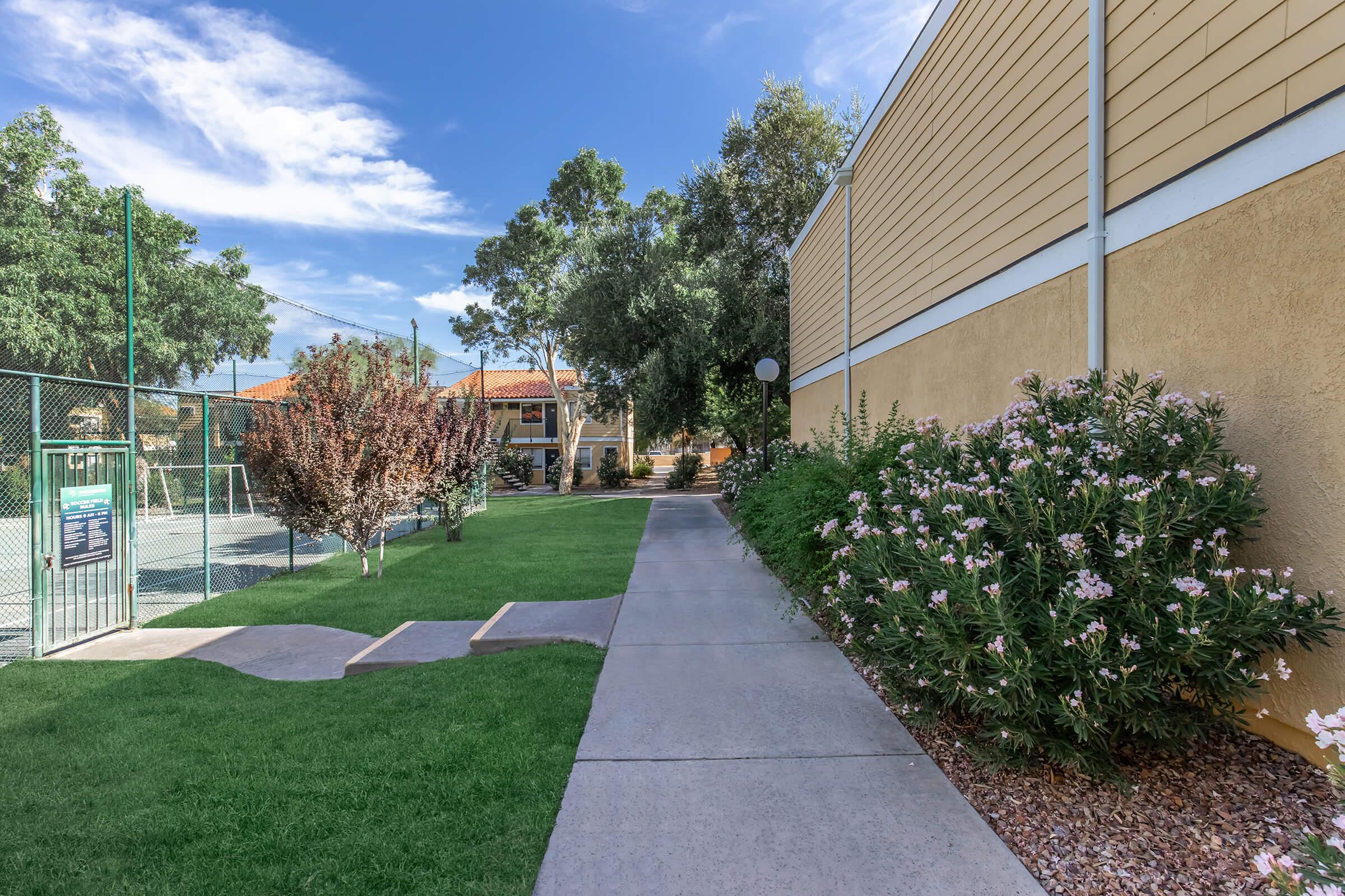 A paved walkway lined with shrubs and grass, leading towards a residential area with tennis courts visible in the background. The scene is bright and sunny, featuring trees and a clear blue sky.