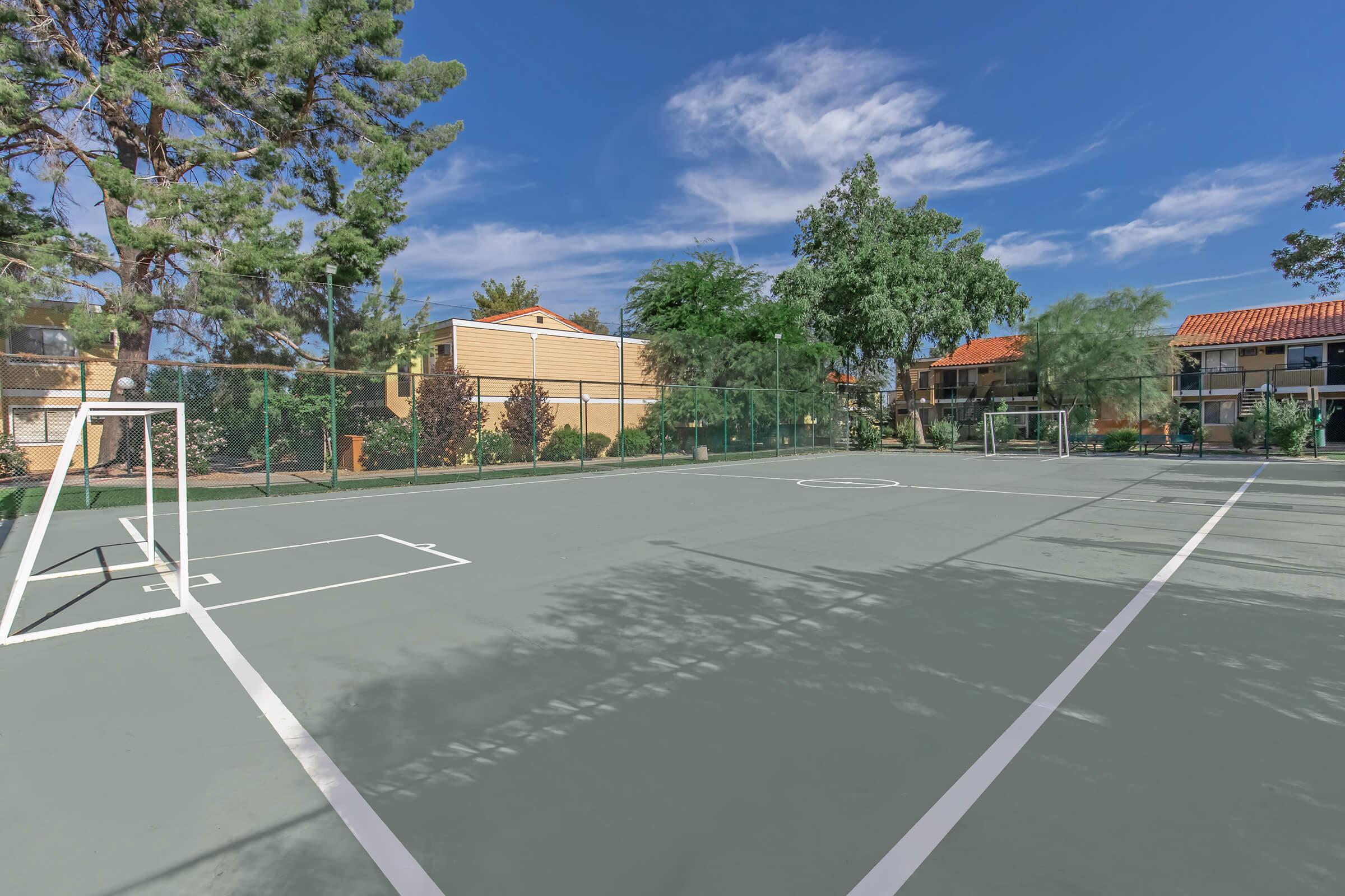 A clear outdoor sports court with two white goalposts on each side. The court is surrounded by trees and buildings, with a blue sky and scattered clouds overhead. The surface is gray, and the area appears well-maintained and ready for use, ideal for playing sports like basketball or soccer.