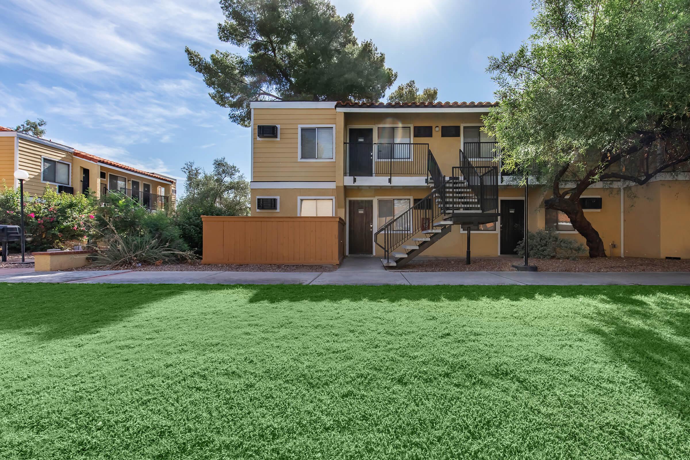 A two-story apartment complex with beige exterior and black staircases, surrounded by lush greenery. In the foreground, there is a well-maintained artificial lawn. The background features additional buildings and trees under a clear sky.