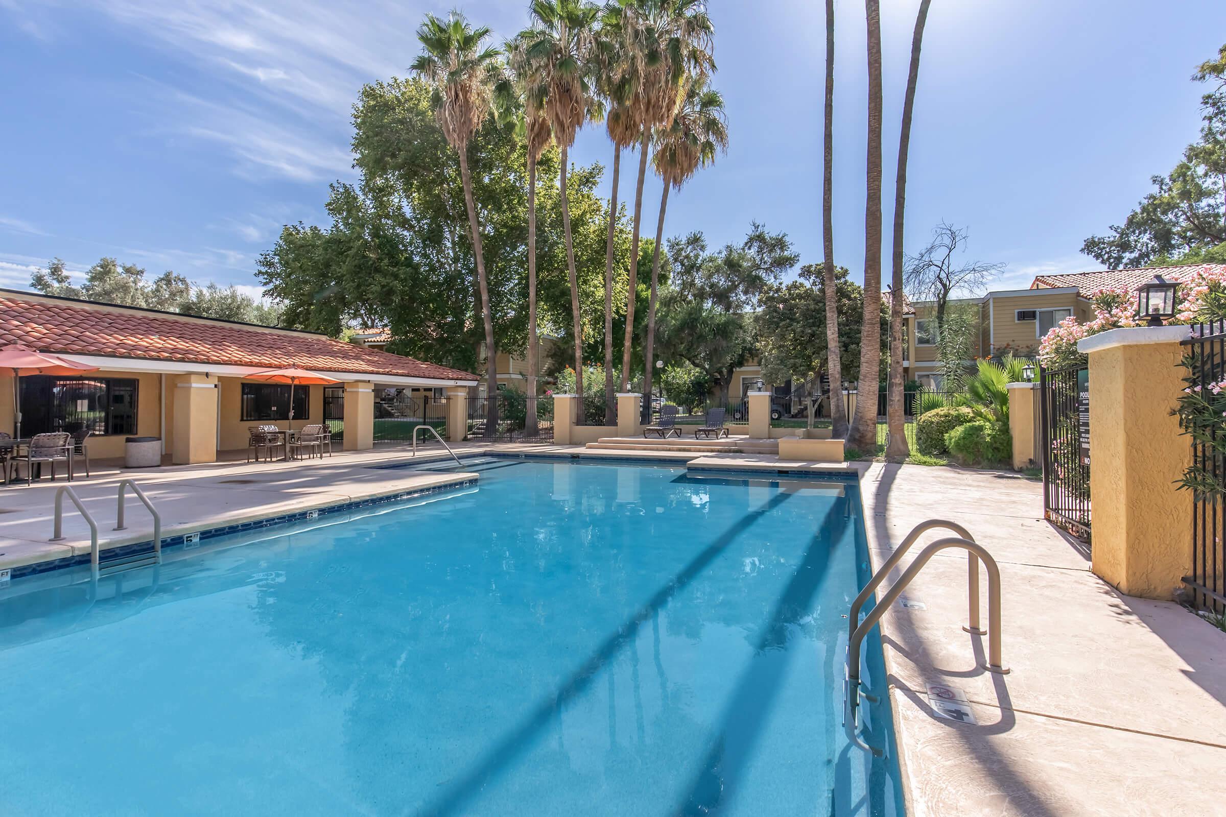 A clear blue swimming pool surrounded by palm trees and lush greenery. Sun loungers and tables are set up nearby, with a building featuring a red-tiled roof in the background. The scene conveys a relaxing outdoor atmosphere.