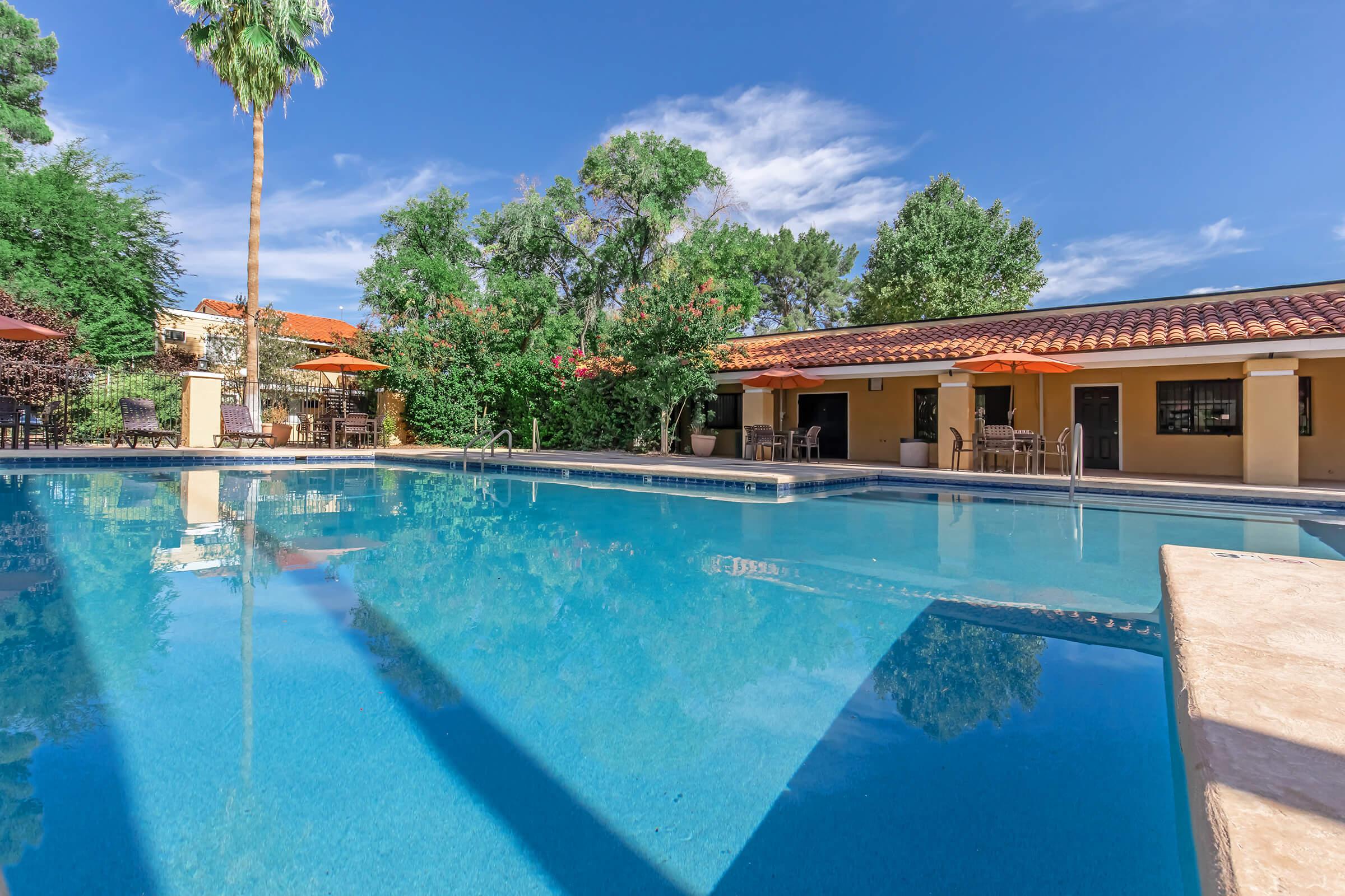 A serene outdoor swimming pool surrounded by lush trees and vibrant landscaping. The pool features clear blue water, with lounge chairs and umbrellas on the deck. In the background, a yellow building with large windows and a tile roof complements the tranquil setting under a clear blue sky.