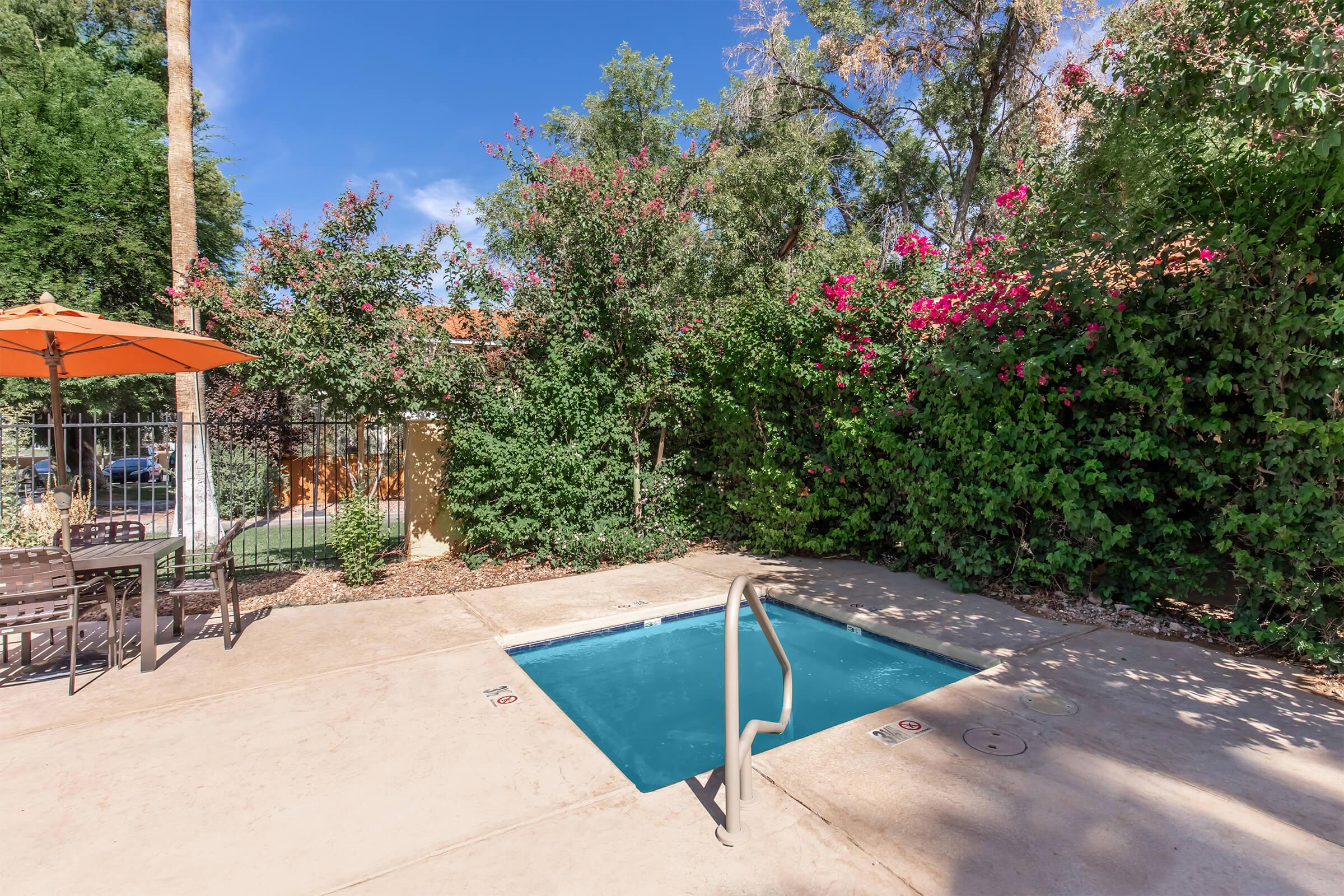 A small private swimming pool surrounded by lush greenery and blooming flowers. There are outdoor seating and an orange umbrella nearby, creating a relaxing atmosphere. The pool area is well-maintained, offering a tranquil retreat. Bright blue sky adds to the inviting ambiance.