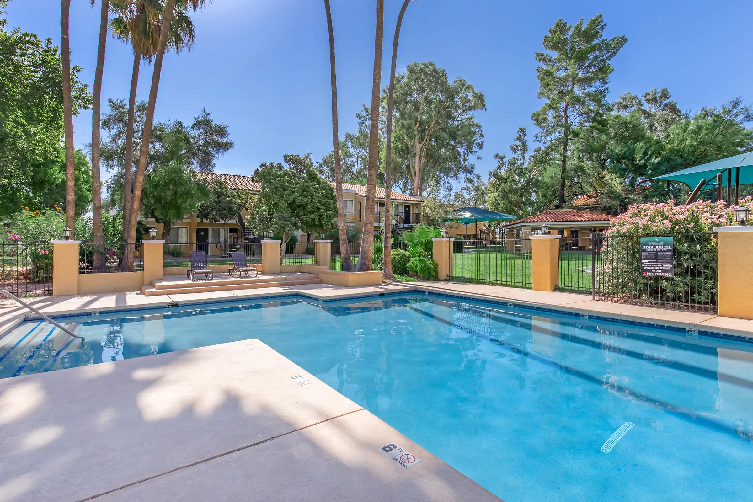 A sunny pool area surrounded by lush greenery, palm trees, and colorful flowers. The pool features a clean, inviting blue water surface with a shallow end visible. Lounge chairs are placed nearby, and there are shaded seating areas under umbrellas, creating a relaxing outdoor atmosphere.