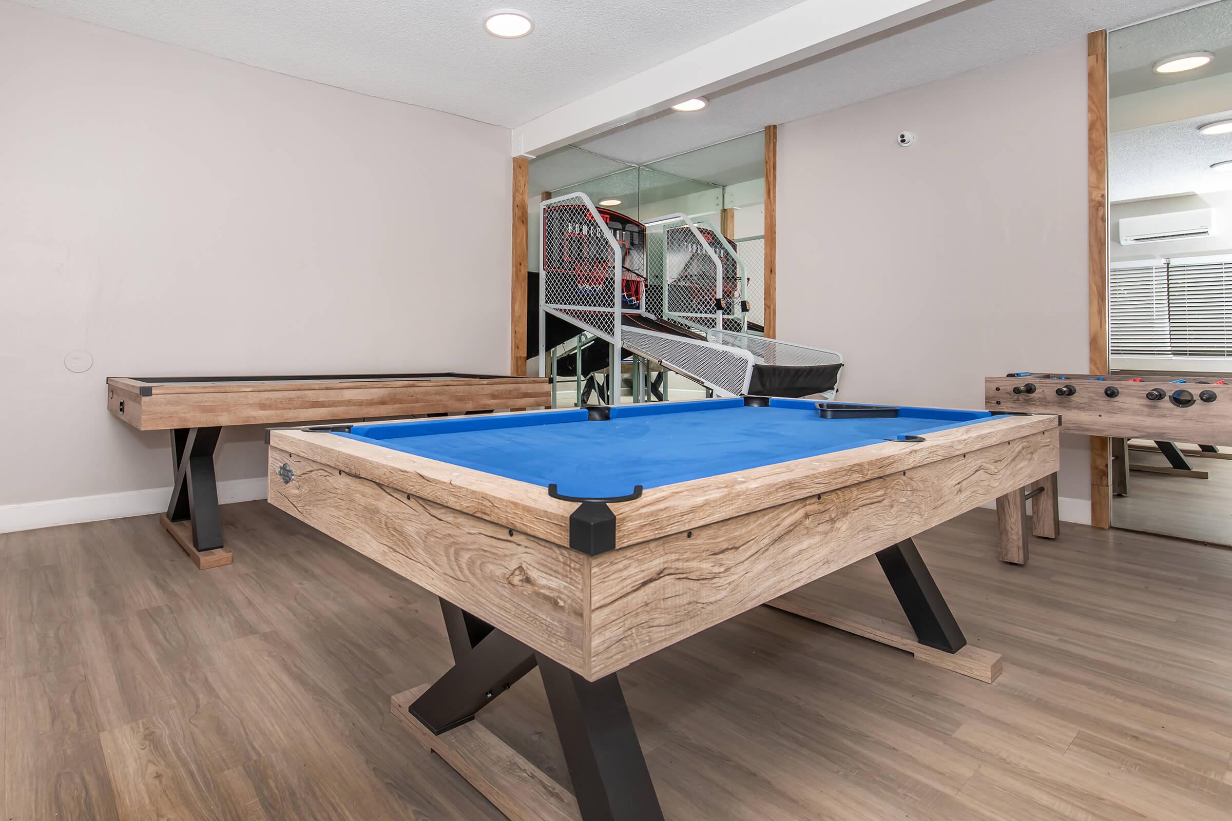 A game room featuring a blue top air hockey table with wooden legs, a foosball table, and a row of modern ramps in the background. The walls are light-colored, and the flooring is wooden, creating a cozy recreational space. Bright overhead lighting enhances the inviting atmosphere.