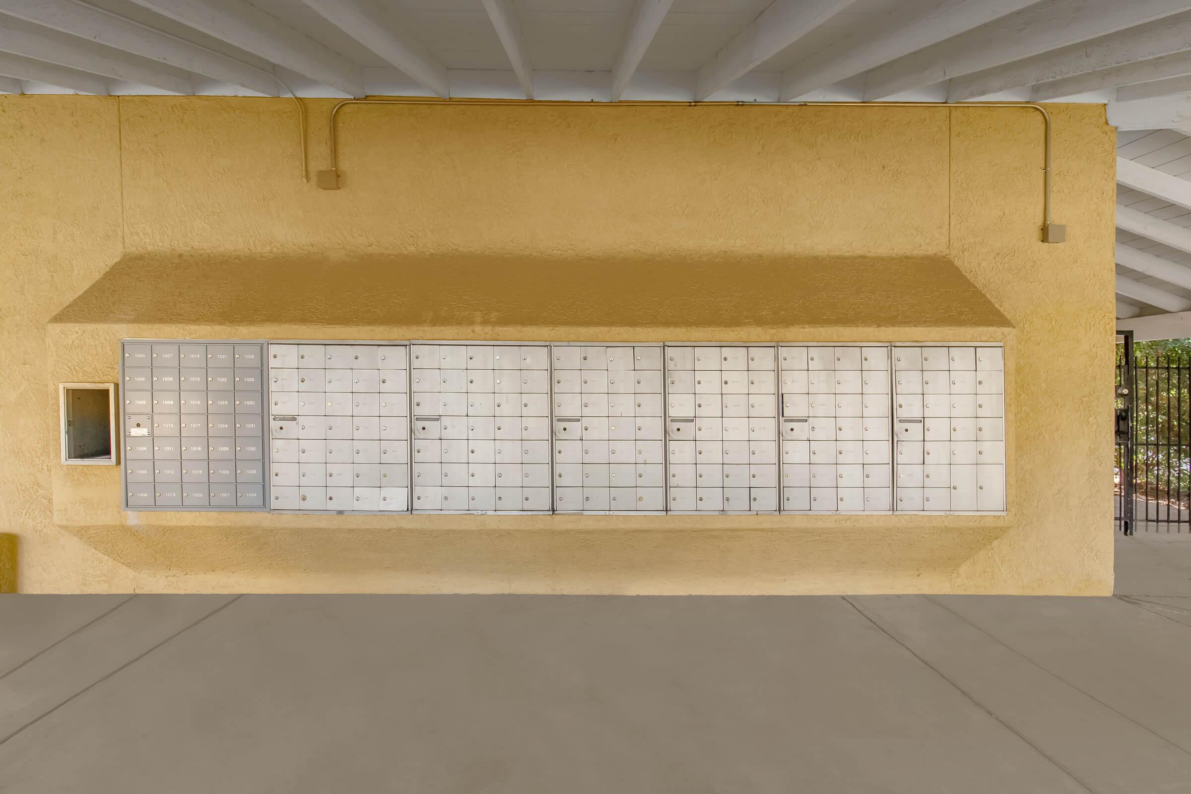 A row of metal mailboxes mounted on a textured yellow wall. The mailboxes are in multiple rows with a small window on the left, and the ceiling features exposed beams. The floor is smooth and light-colored.