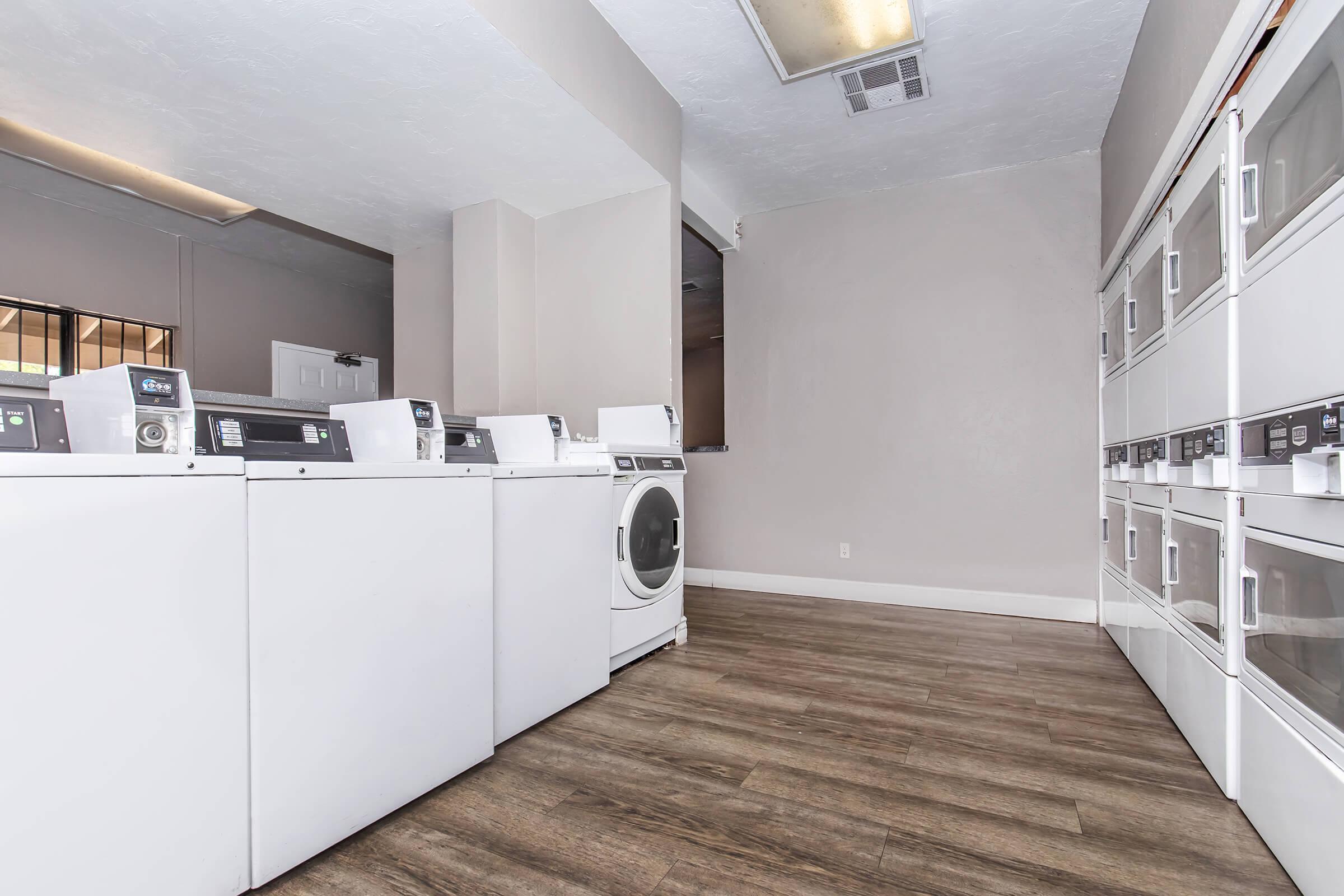 A clean and modern laundry room featuring several white washing machines and a dryer. The floor is made of wood-like vinyl planks, and the walls are painted in a neutral color. There is a door visible in the background and a countertop area next to the appliances.