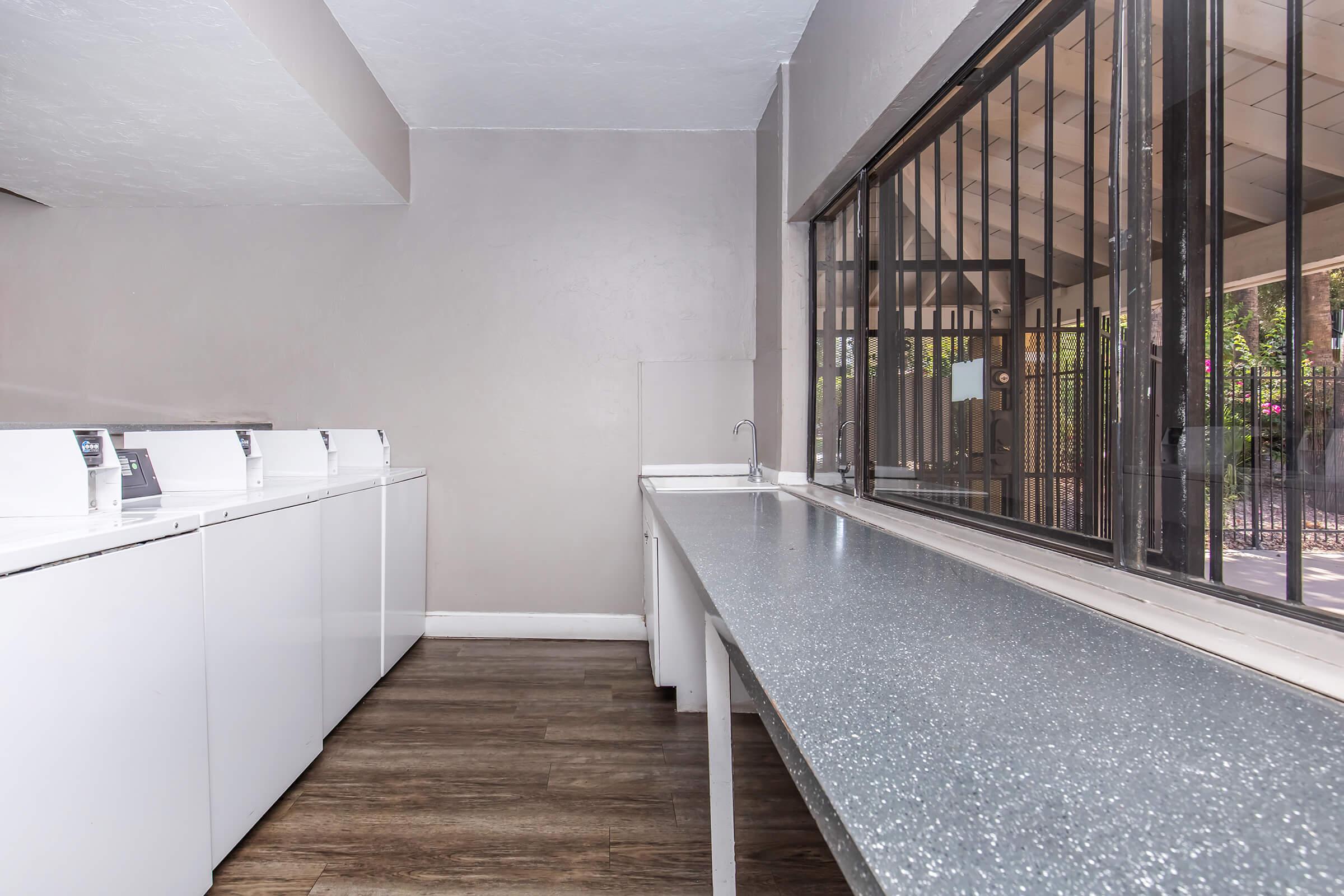 A clean, modern laundry room featuring several white washing machines lined against one wall. A long gray countertop runs alongside the machines, with a window allowing natural light and a view outside. The space is well-lit and has a simple, minimalist design.