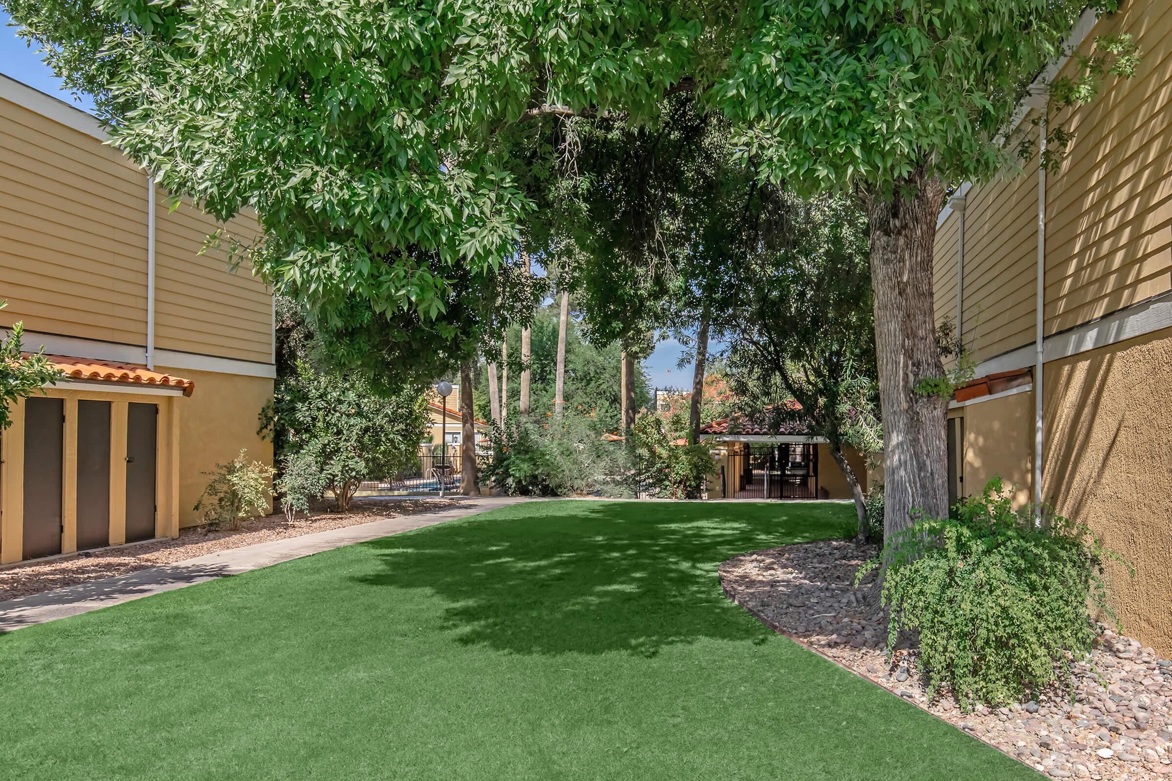 A lush green artificial lawn surrounded by trees and landscaped paths, with nearby buildings featuring yellow siding. Sunlight filters through the foliage, creating a serene outdoor space.