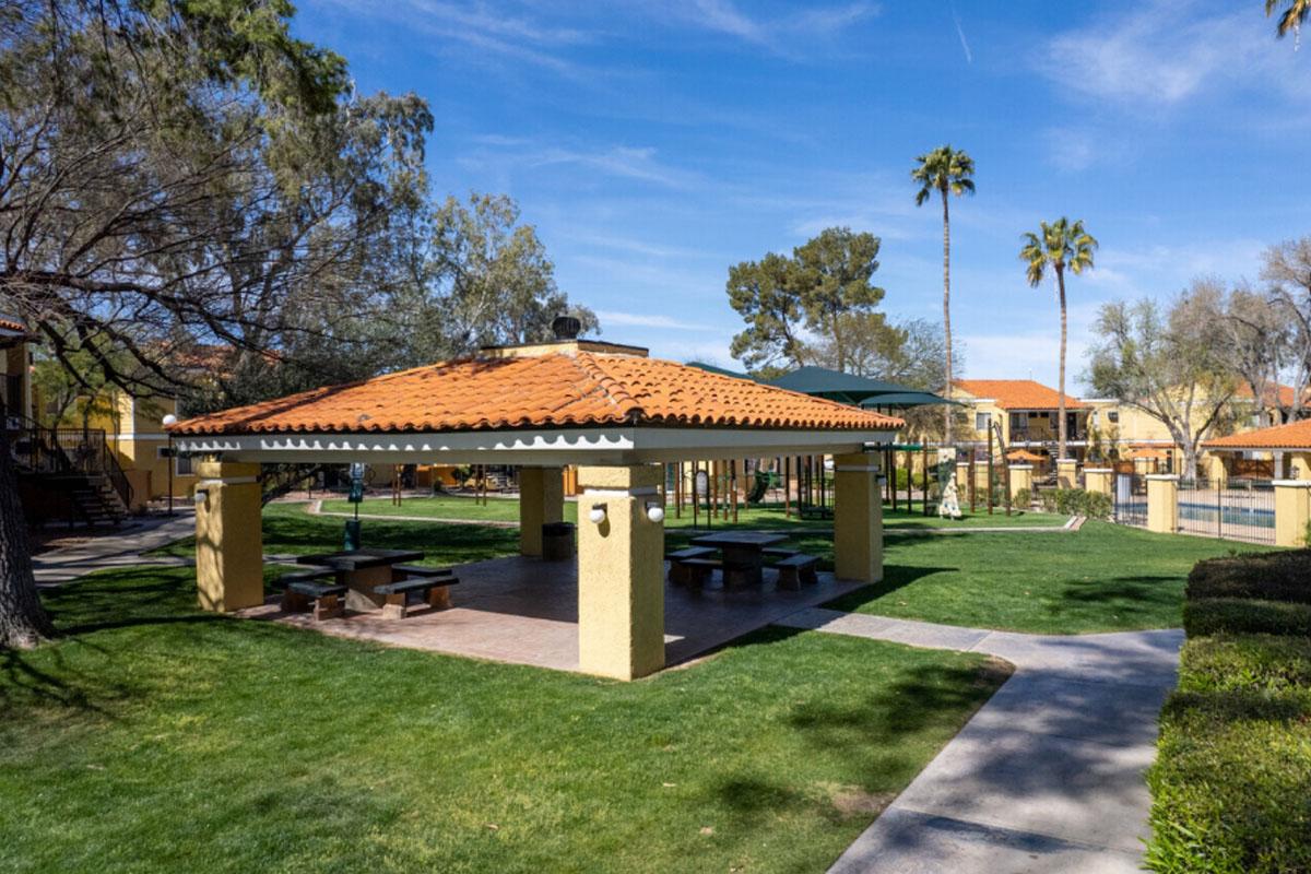 A sunny park scene featuring a large gazebo with a red tiled roof. Surrounding the gazebo are green lawns and palm trees, with playground equipment in the background. The sky is clear with a few clouds, creating a vibrant outdoor atmosphere.