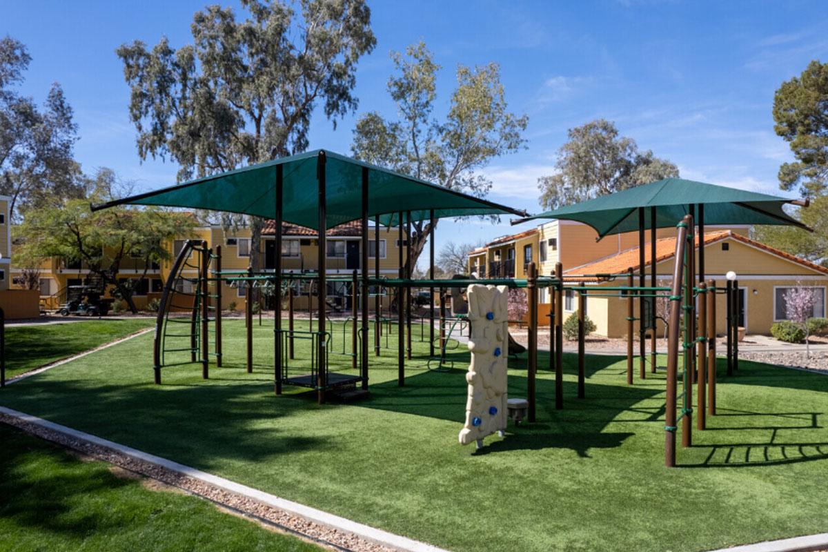 A playground featuring green turf, two large shaded areas with canopies, climbing structures, and a rock wall. Surrounding the playground are trees and residential buildings in the background, creating a lively outdoor space for children to play.