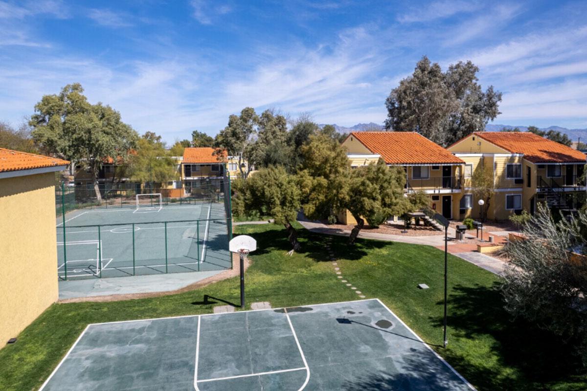 A view of a residential complex featuring a basketball court in the foreground and several buildings in the background. The court is surrounded by greenery and trees, with clear blue skies above. The scene captures a serene outdoor environment ideal for recreation and community activities.