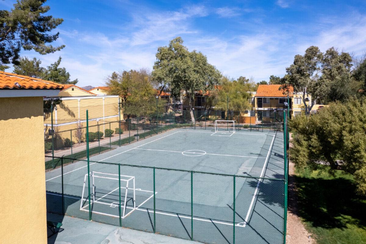 Aerial view of a basketball court surrounded by trees and residential buildings. The court has two goalposts at each end, a central circle, and is enclosed by a fence. Clear blue sky and scattered clouds provide a bright atmosphere.