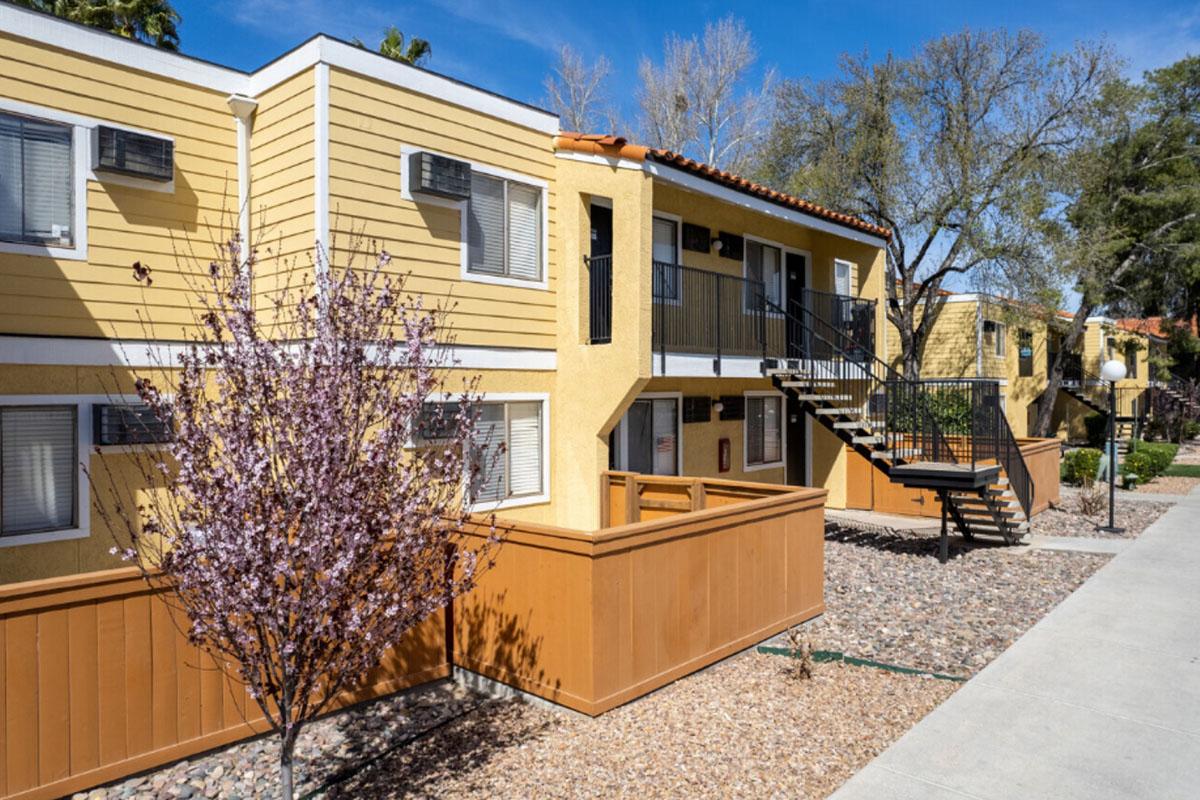 Row of yellow apartment buildings with a staircase and small trees in front. The courtyard features decorative gravel and landscaping. There are windows with air conditioning units, and the sky is clear with a few scattered clouds.