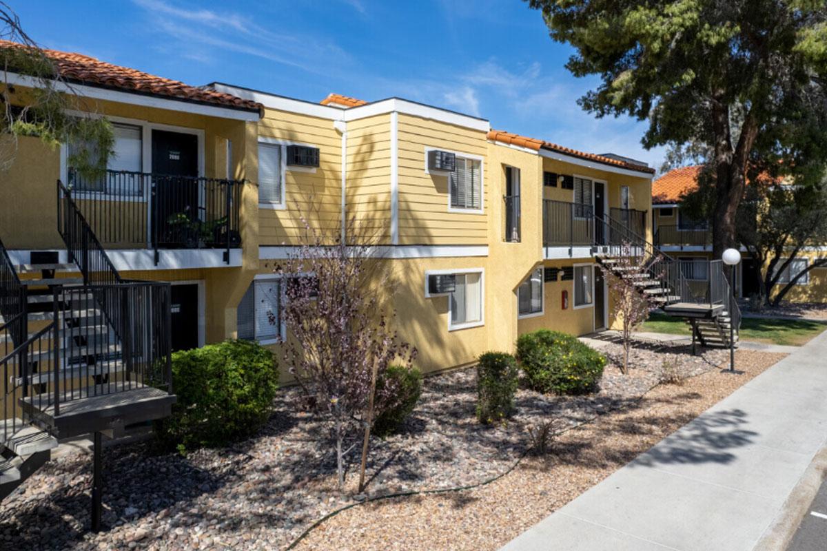 A view of a two-story residential apartment complex with yellow exterior walls, black railings, and balconies. There are small bushes and gravel landscaping in front, with a clear blue sky above. The scene is bright and well-maintained, typical of suburban living.