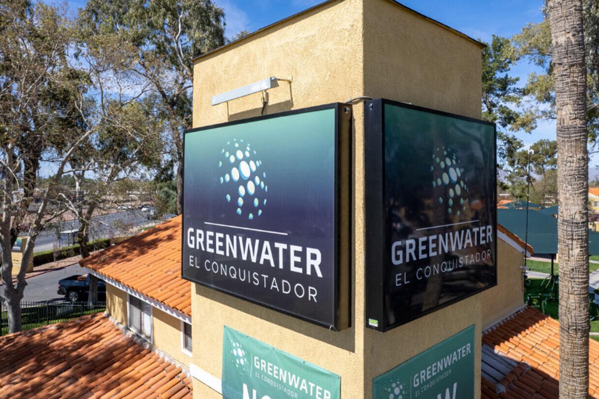 Signage for Greenwater El Conquistador displayed on a building, featuring a modern logo and the name prominently in bold text. The background includes trees and a blue sky, indicating an outdoor setting. The overall design reflects a professional and inviting atmosphere.