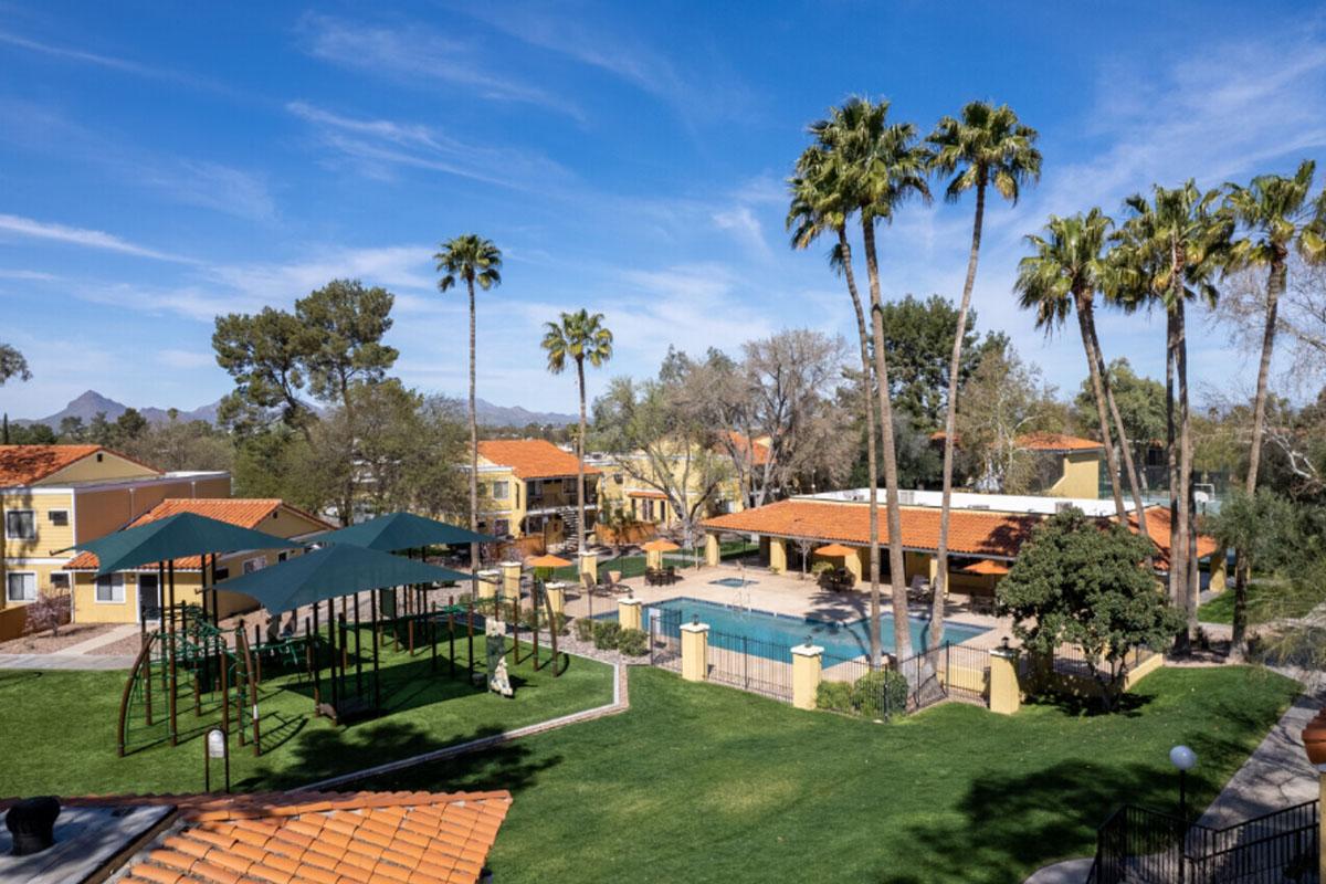 Aerial view of a residential area featuring a swimming pool, palm trees, and a playground. Vibrant green grass surrounds the pool and playground, with several colorful buildings and mountain peaks in the background. The sky is clear and blue, creating a sunny, inviting atmosphere.