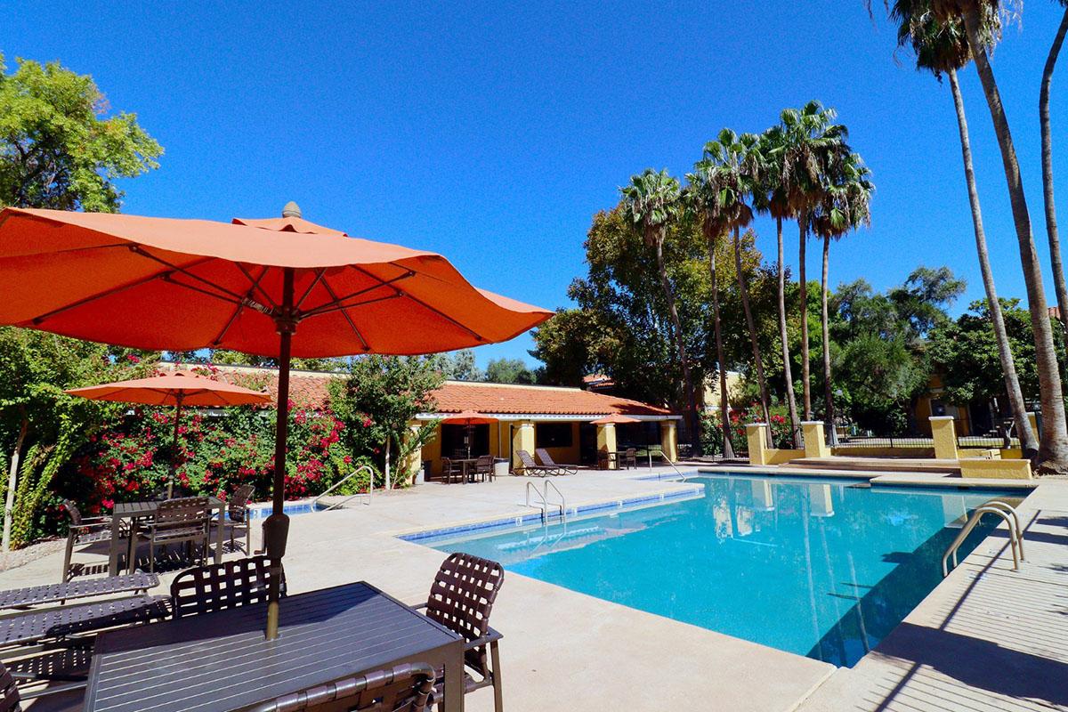 A sunny outdoor pool area featuring a clear blue pool surrounded by lounge chairs and tables with orange umbrellas. Lush greenery and palm trees provide a tropical ambiance, with a building in the background. Ideal for relaxation and leisure activities.