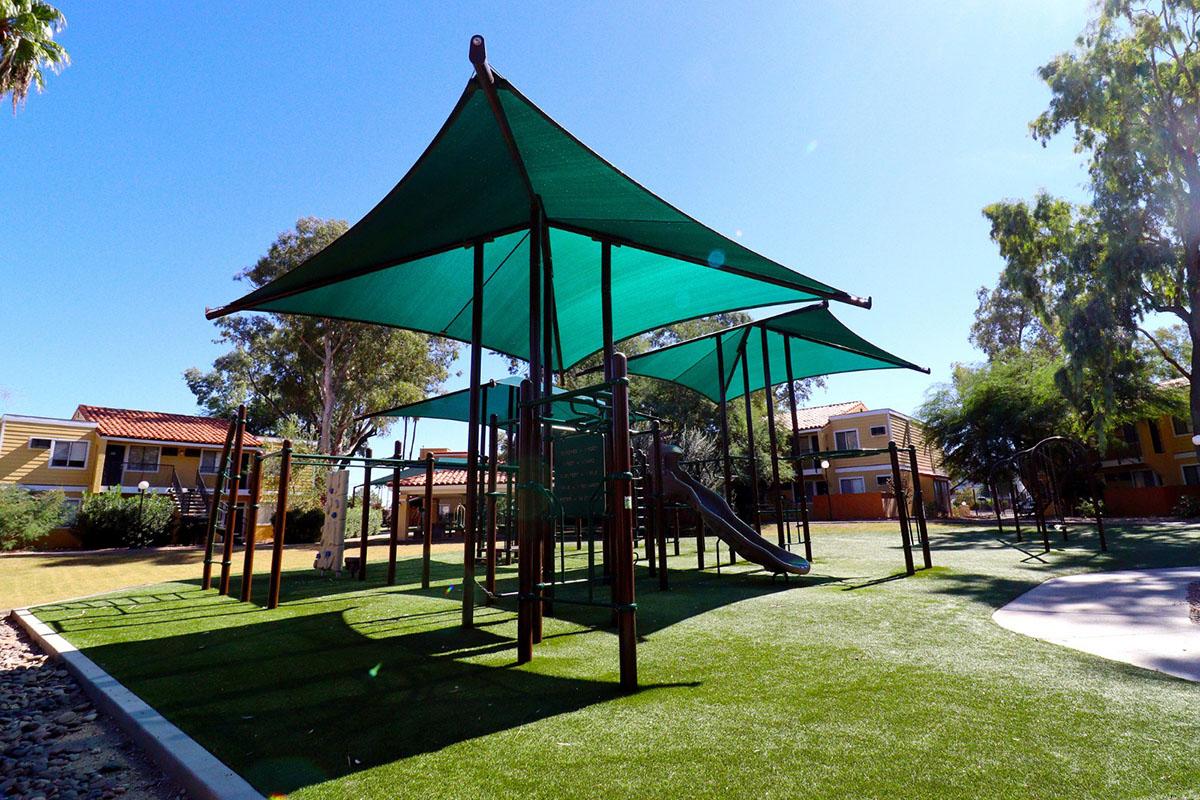 A playground featuring green shade sails, climbing structures, a slide, and artificial turf, surrounded by trees and residential buildings in the background under a clear blue sky.