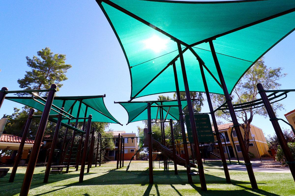A brightly lit playground featuring green shade sails, with metal climbing structures visible under the sunny sky. Trees and apartment buildings are in the background, creating a vibrant and inviting outdoor space for children to play.