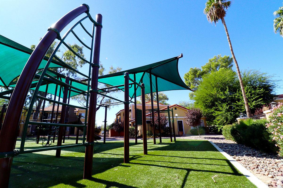 A playground set with climbing structures and shade canopies, situated on artificial turf. Surrounding the playground are palm trees and well-kept landscaping, with residential buildings visible in the background under clear blue skies.