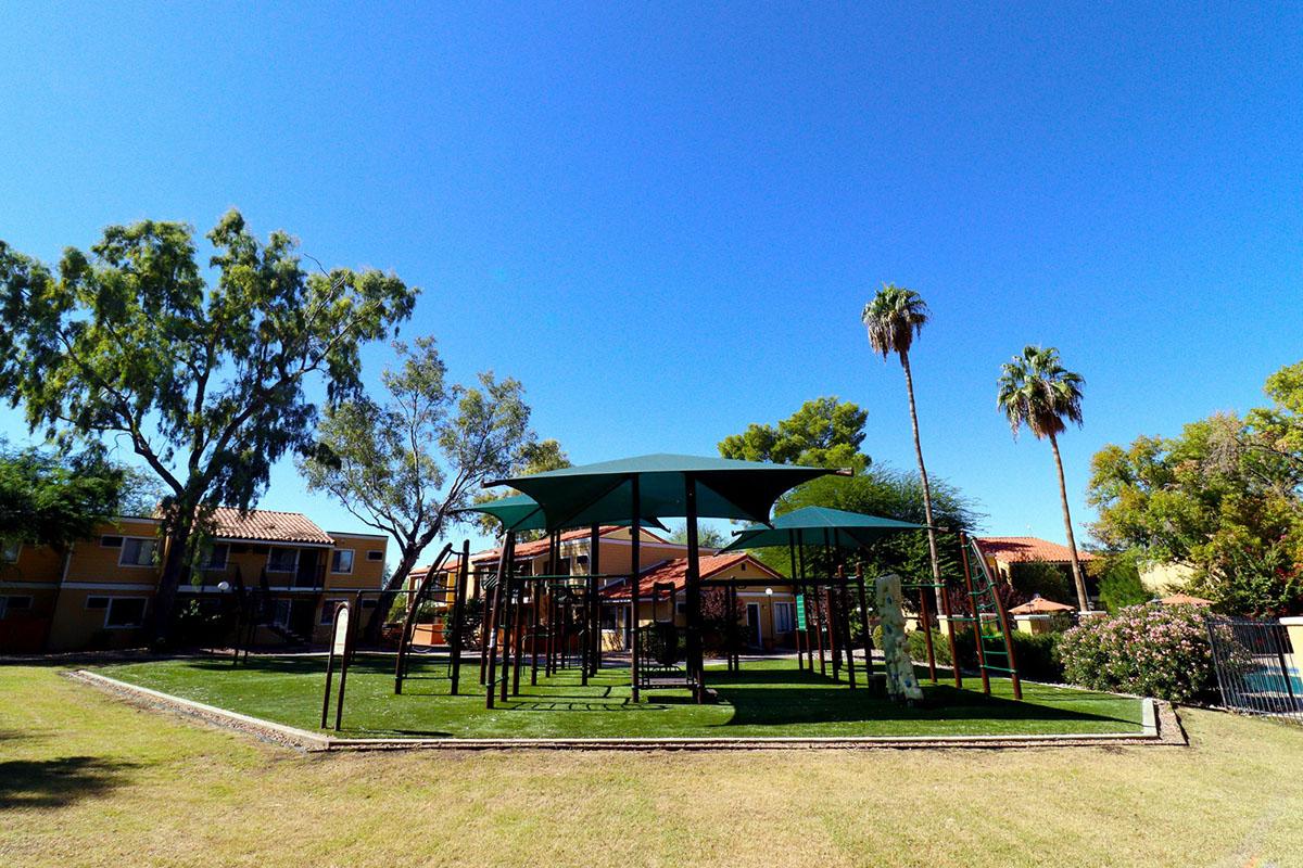A playground featuring green shaded structures and climbing equipment, surrounded by grassy areas. In the background, there are residential buildings with a clear blue sky. Palm trees and other greenery enhance the outdoor setting.