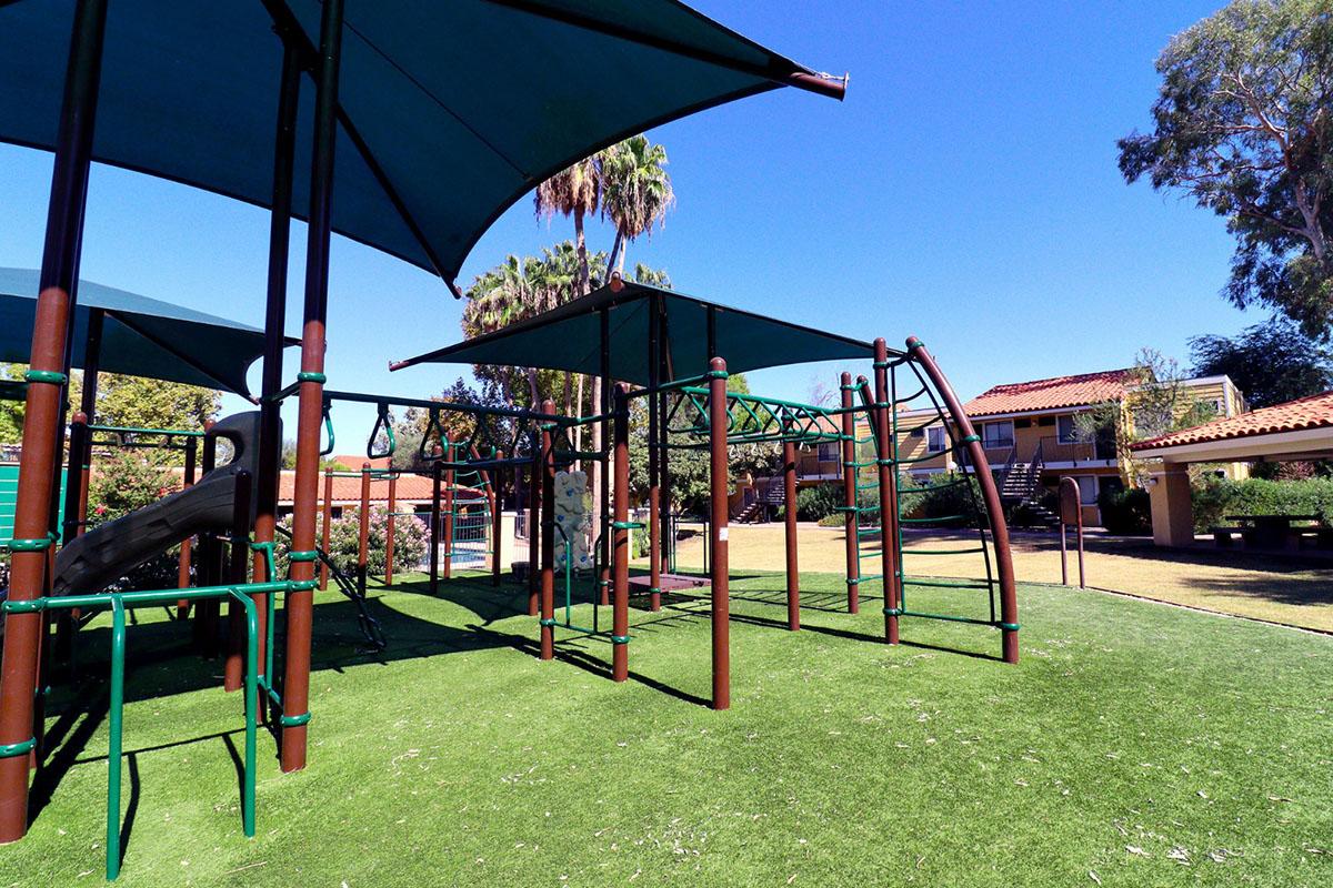 A playground featuring climbing structures, a slide, and shade canopies, set on green grass. In the background, palm trees and a building are visible under a clear blue sky.
