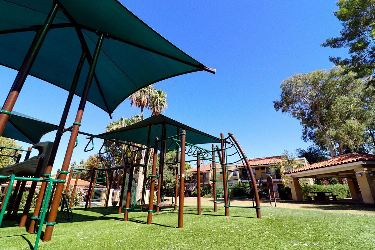 A vibrant playground featuring green turf, shaded areas with large canopies, and climbing structures. In the background, there are residential buildings and palm trees under a clear blue sky, creating a sunny and inviting atmosphere for play and leisure.